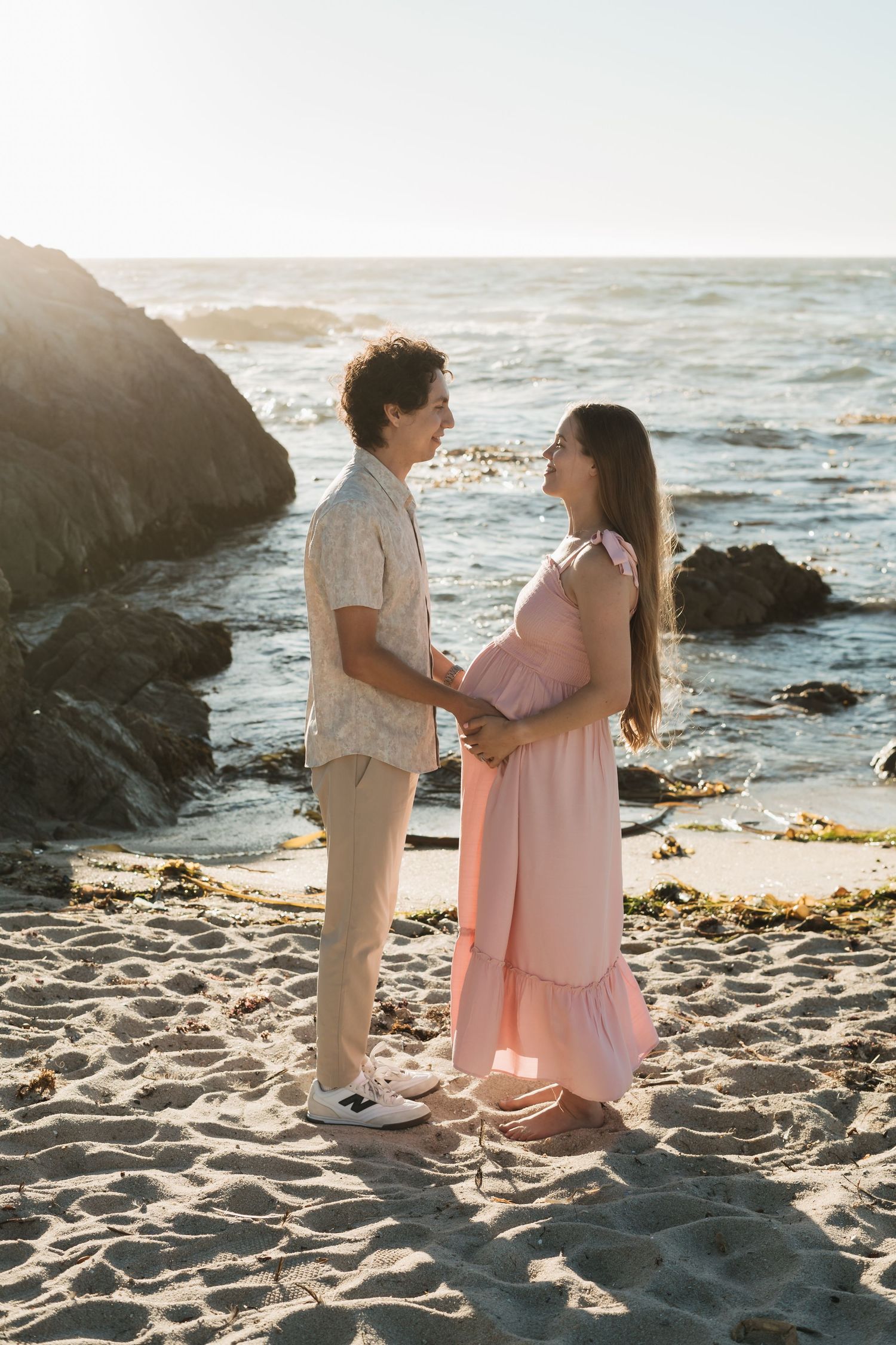 A couple shares a romantic moment on a sandy beach at sunset with ocean waves and rocks in the background.
