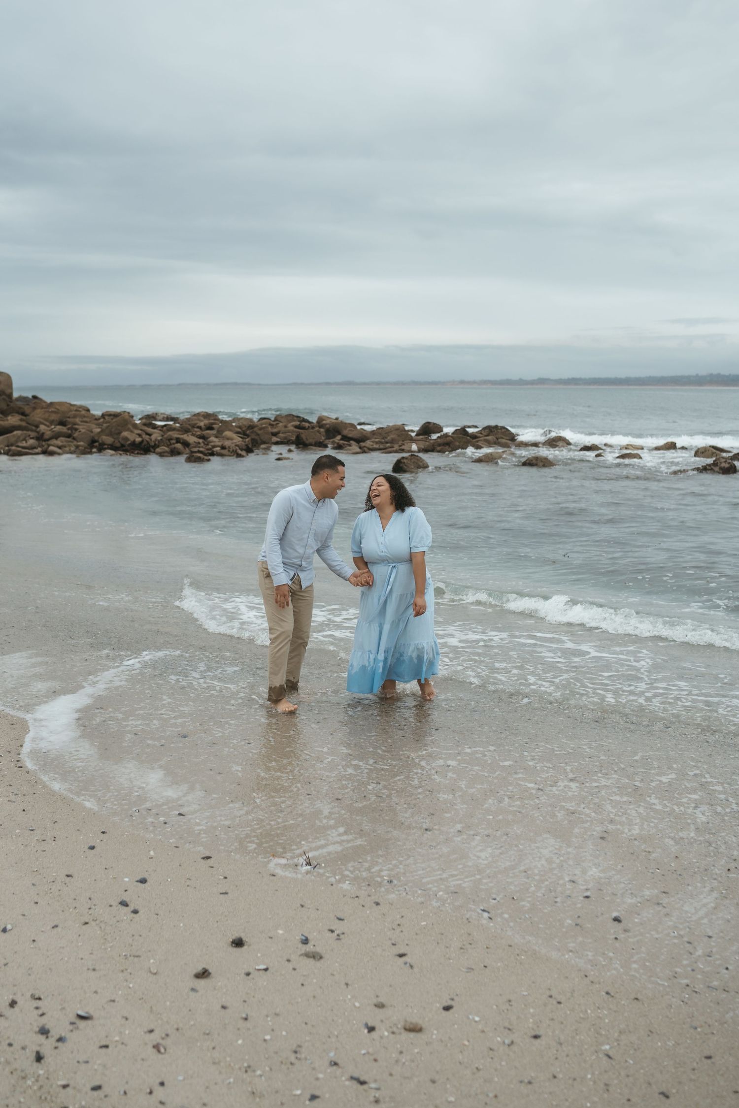 Three people in light colored clothing stand at the edge of the ocean waves on a cloudy day at the beach.