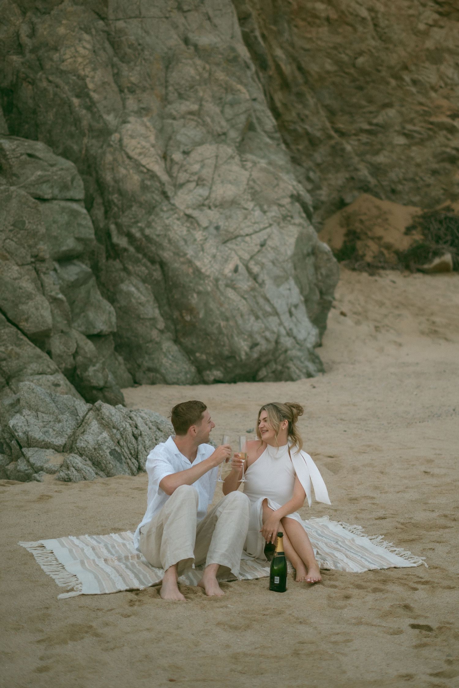 Couple sitting on a blanket on the beach toasting with champagne glasses in front of large rocks.