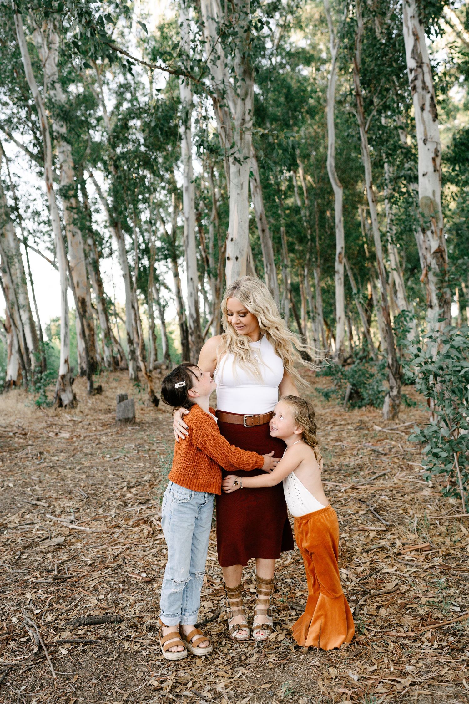 A blonde mother smiles as her two children hug her in a eucalyptus forest.