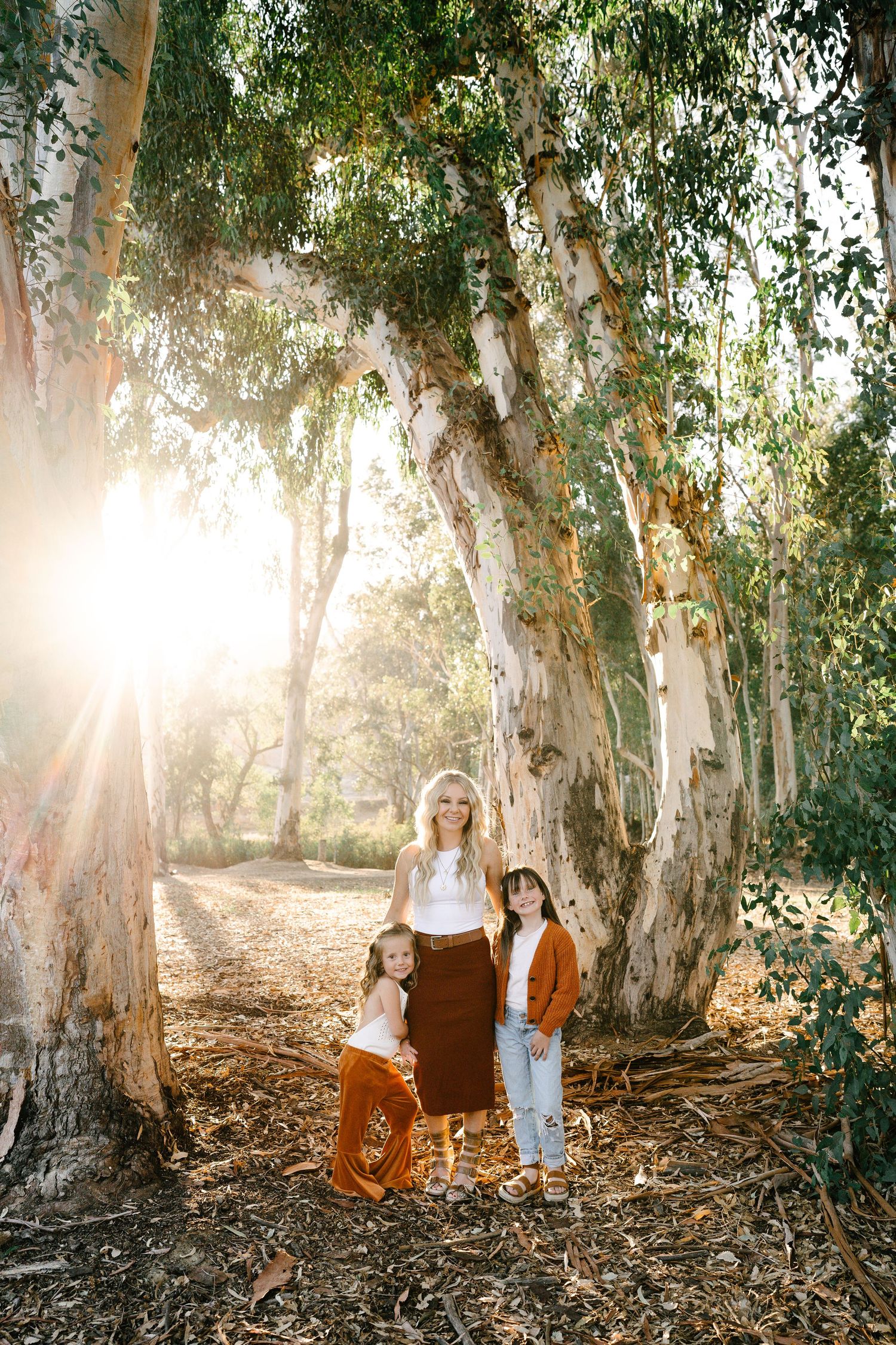 A mother and two children pose among towering eucalyptus trees with golden sunlight streaming through the forest.