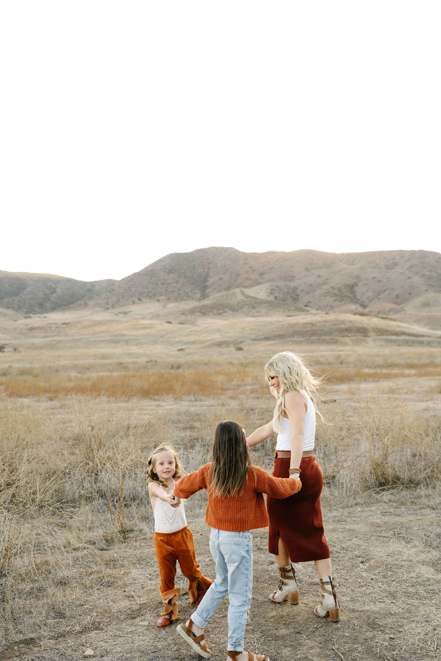 A mother and two children hold hands dancing in a golden field with rolling hills in the background.