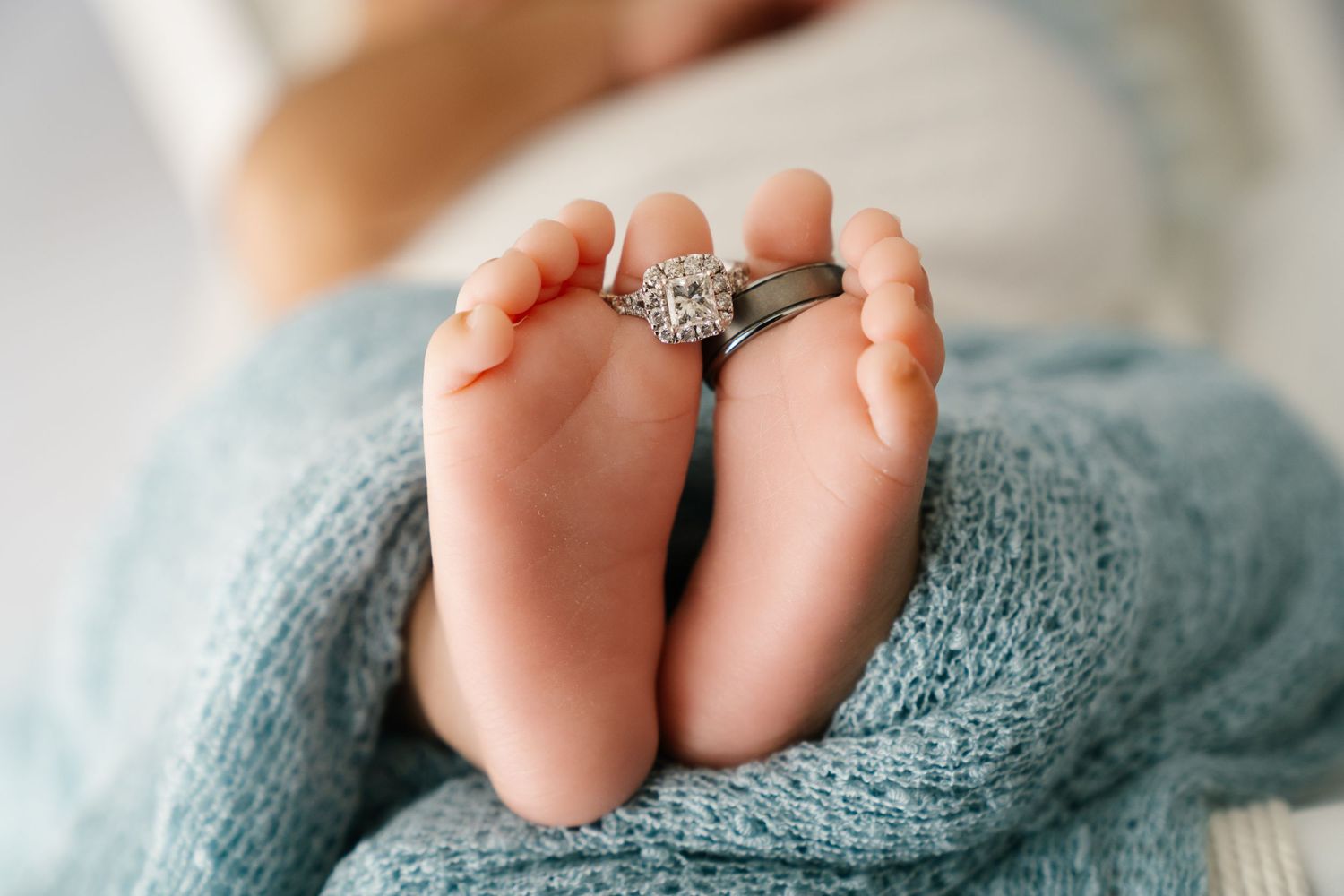 Tiny newborn feet with engagement ring placed between them on blue knit blanket.