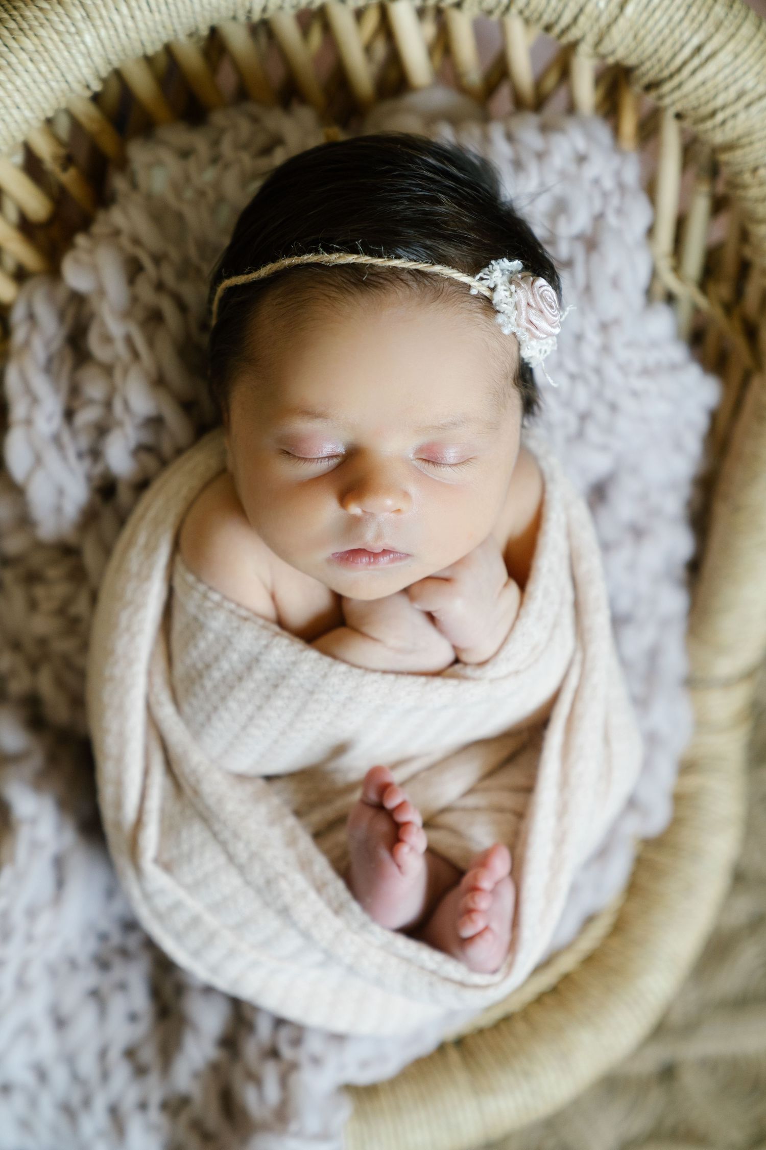 Sleeping newborn wrapped in cream fabric with delicate headband in woven basket.