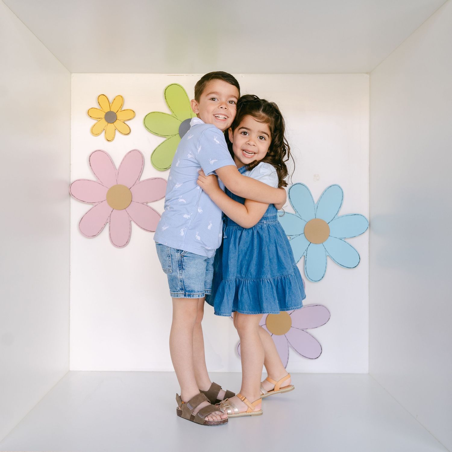 Two young children wearing blue embrace in front of a wall decorated with colorful pastel flower designs.