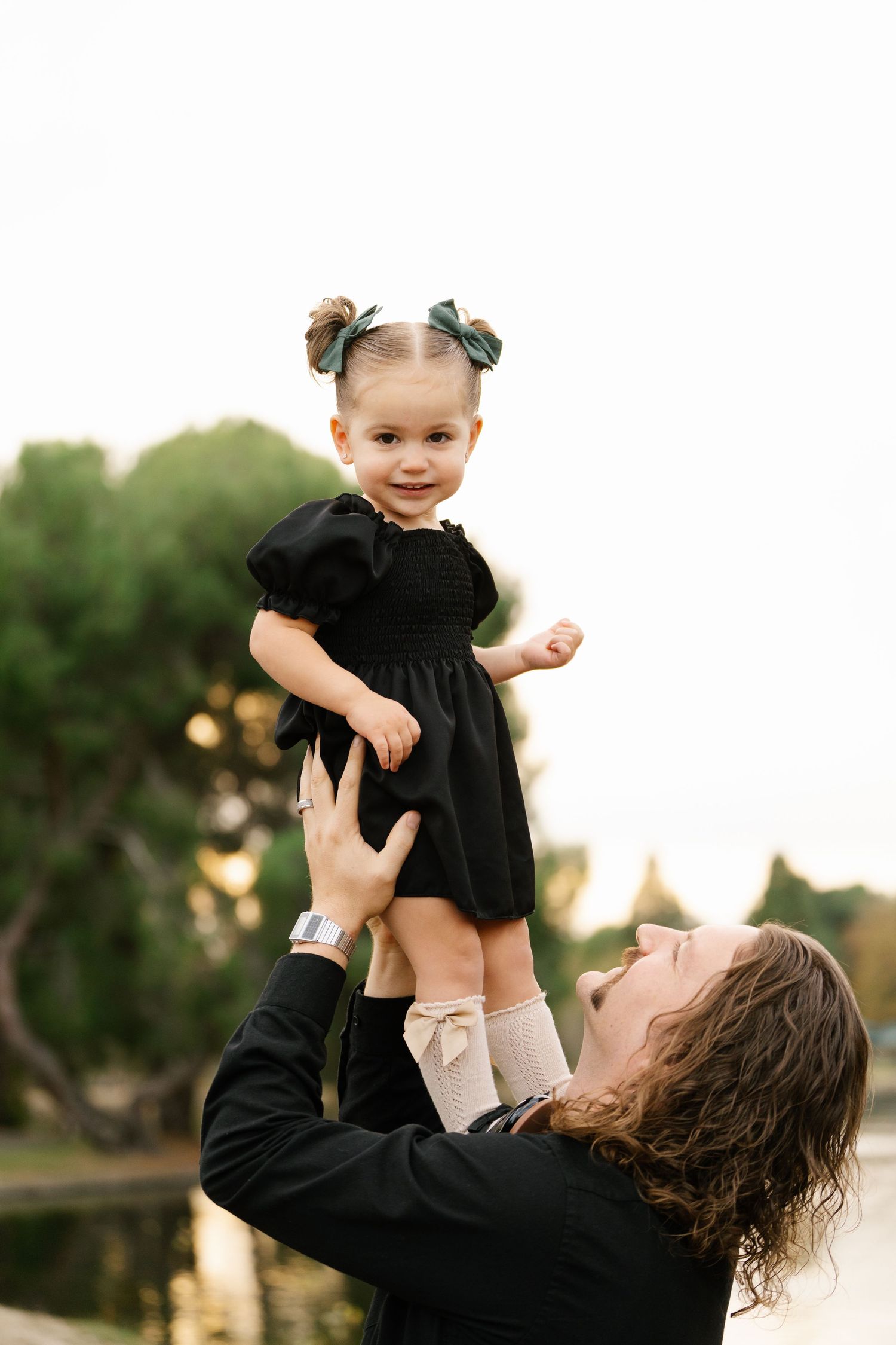 Small child in black dress being lifted up high against nature backdrop.