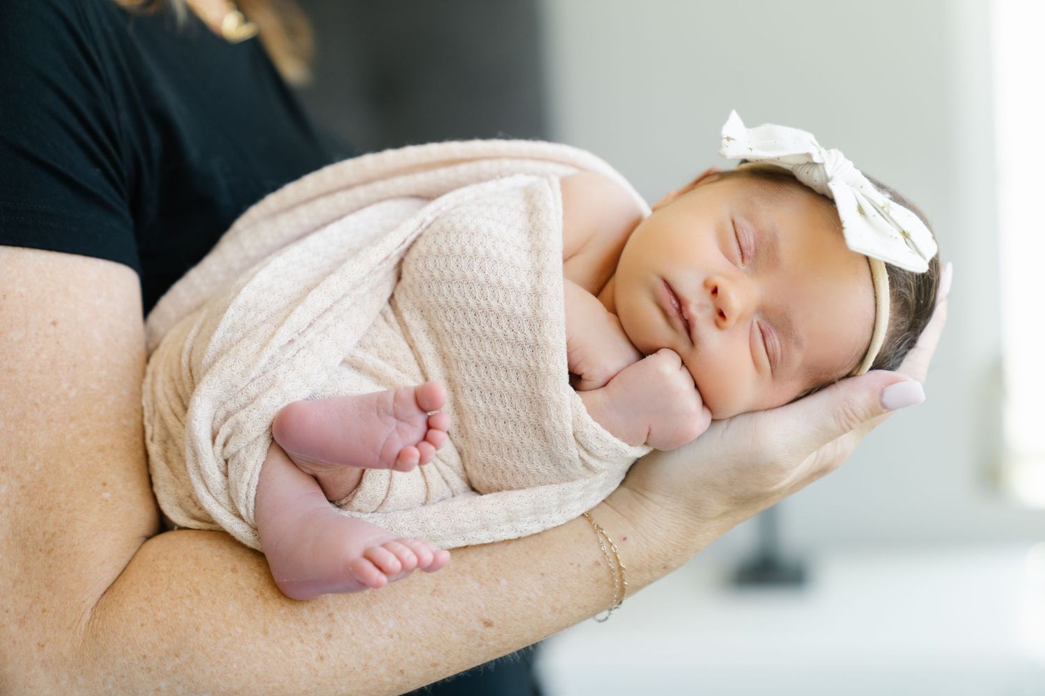 Sleeping newborn in cream wrap with bow accessory being cradled.