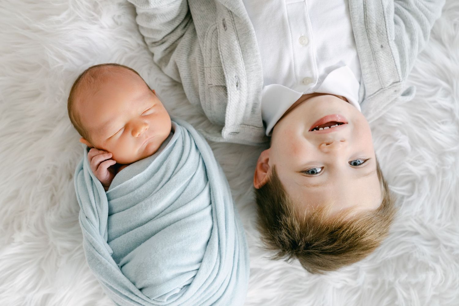 Two sleeping infants lying on white furry blanket, one swaddled in blue.