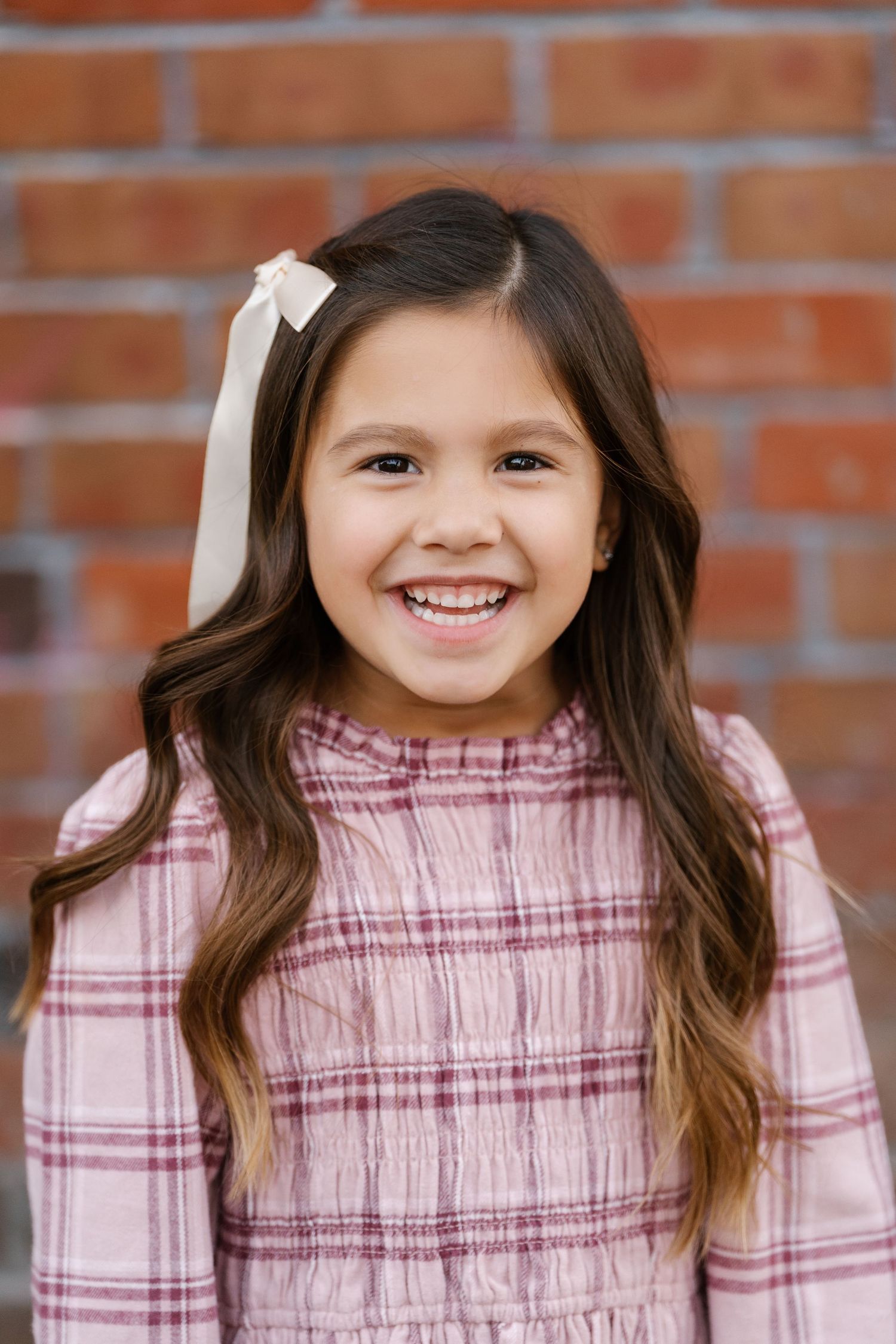 A young child in a pink plaid dress smiles against a brick wall background.