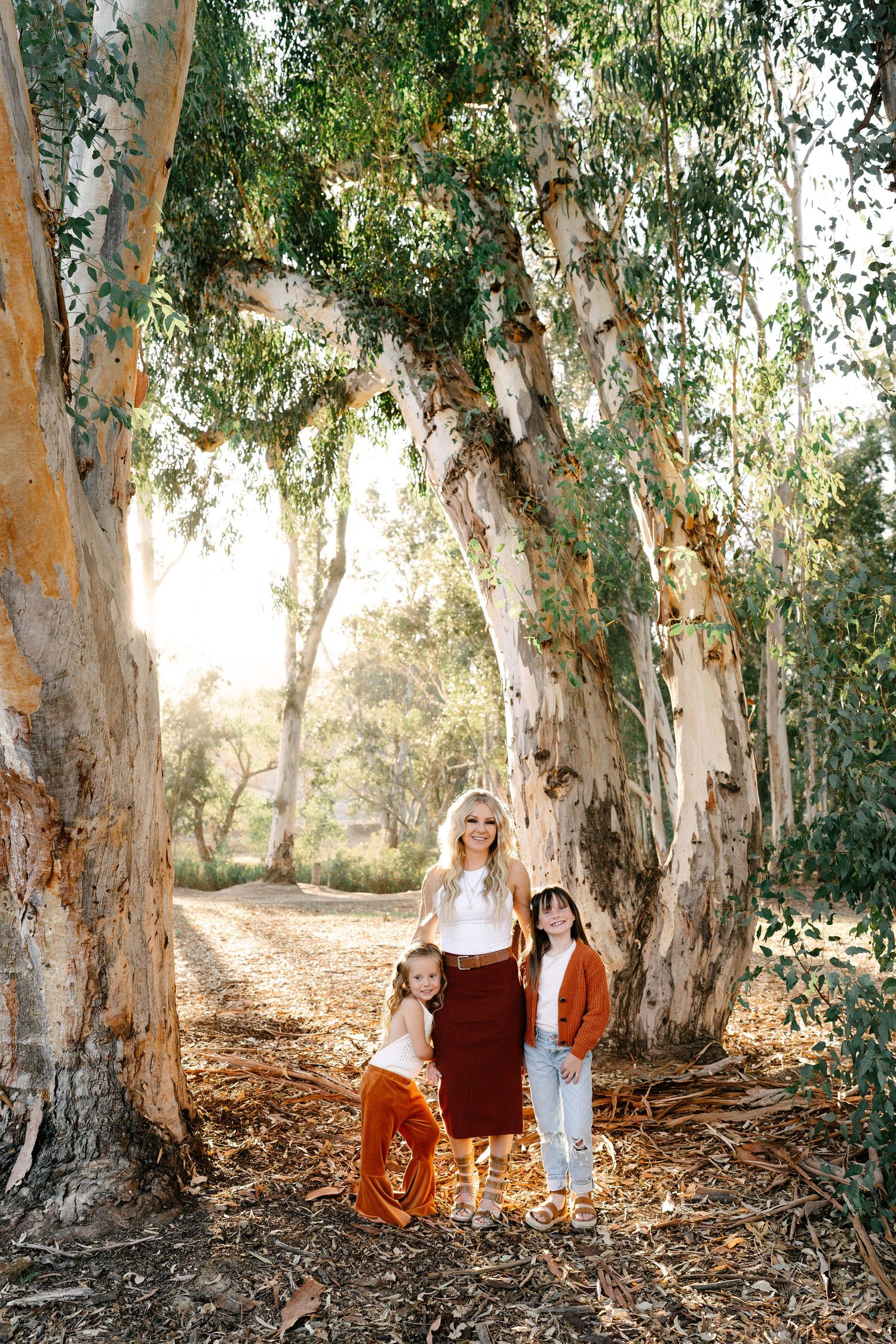 A blonde woman poses with two young girls under large eucalyptus trees in a sunlit forest path.