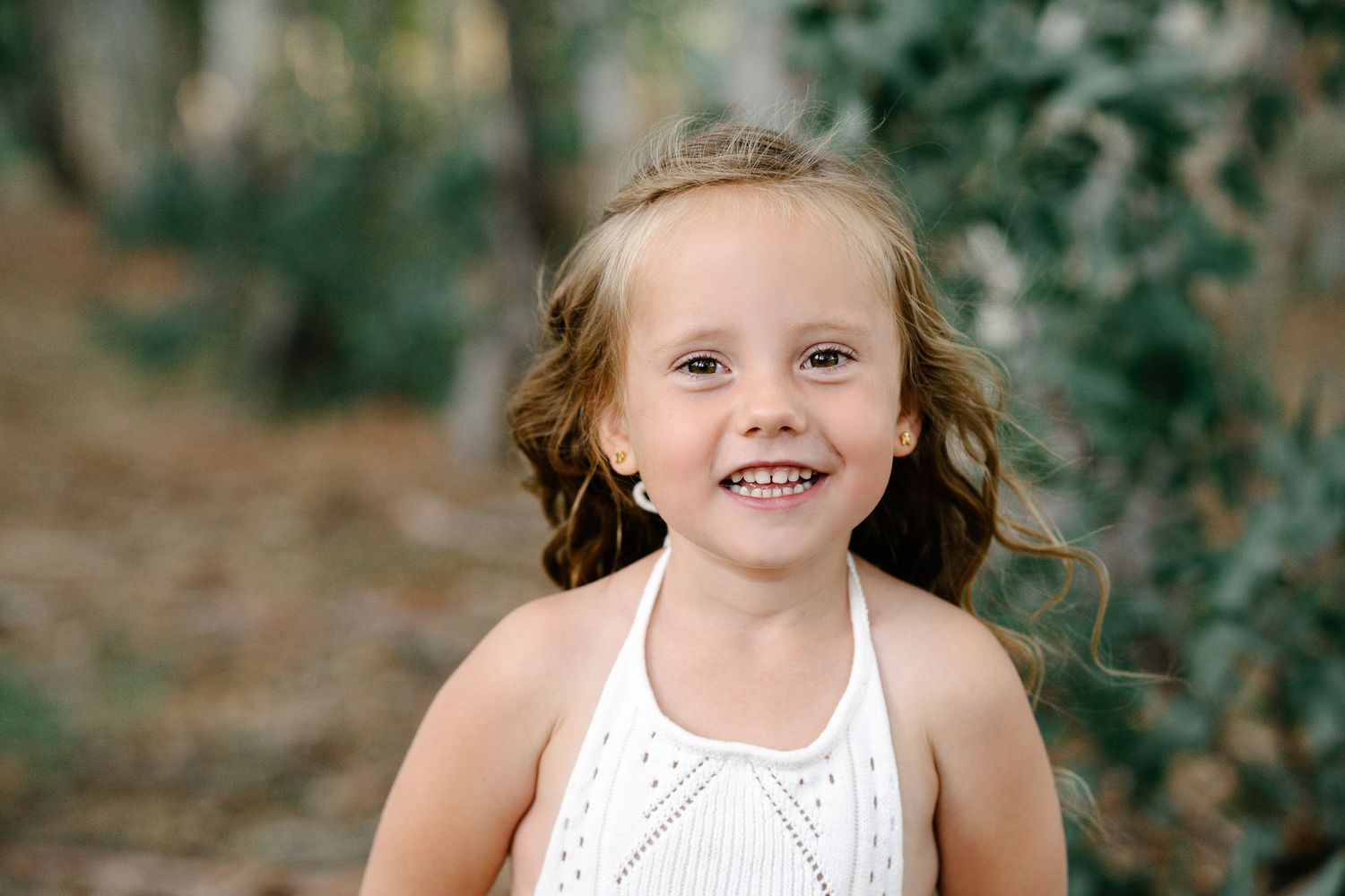 A smiling young girl with curly hair wearing a white dress poses outdoors among green trees.