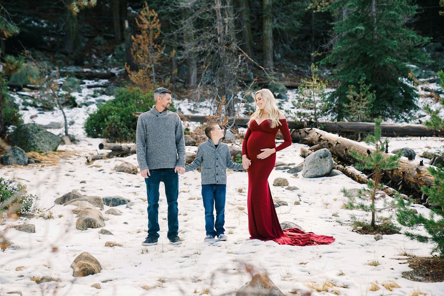 Winter maternity photo of family standing in snowy forest landscape.