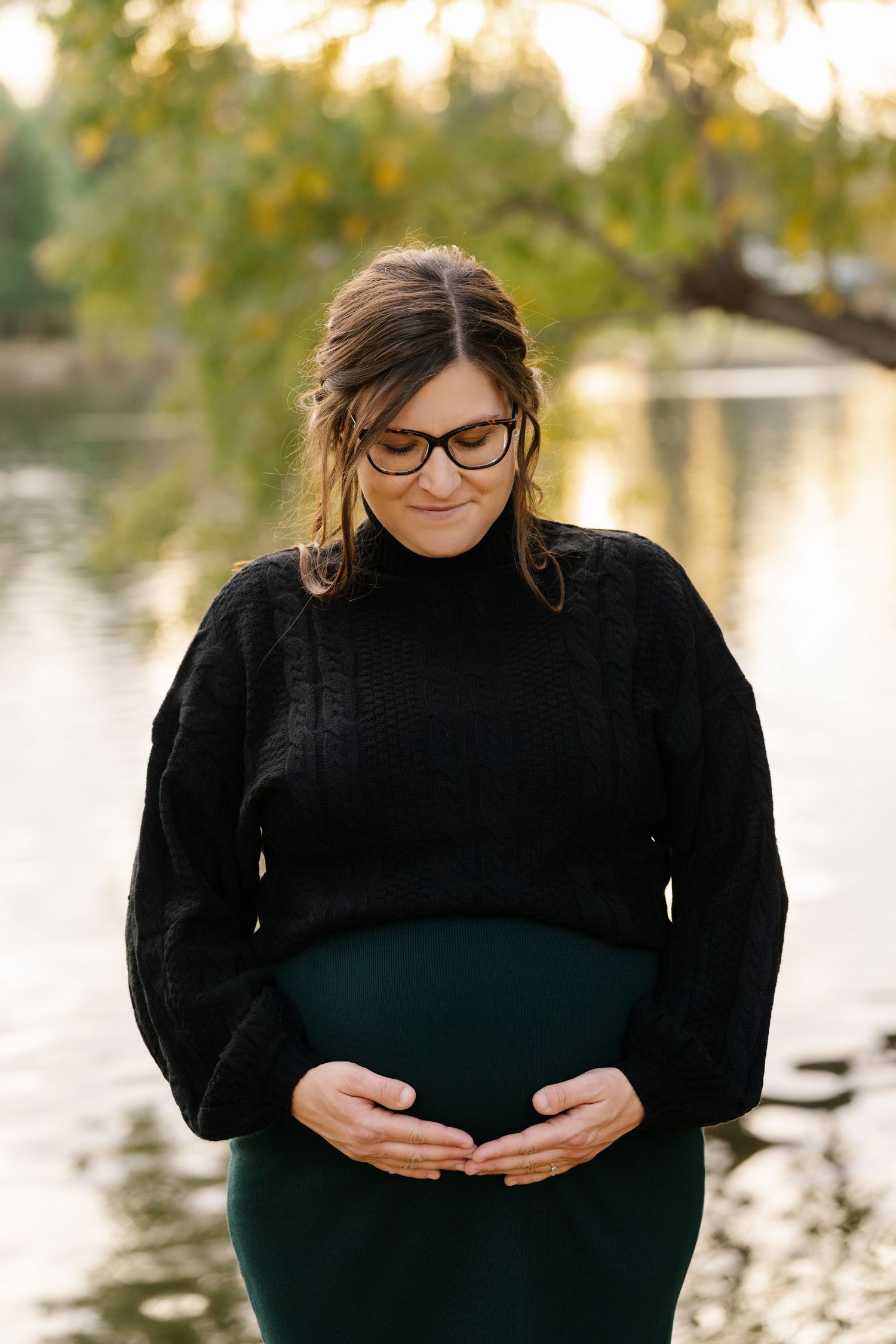An expectant mother in a black sweater stands near a peaceful pond.