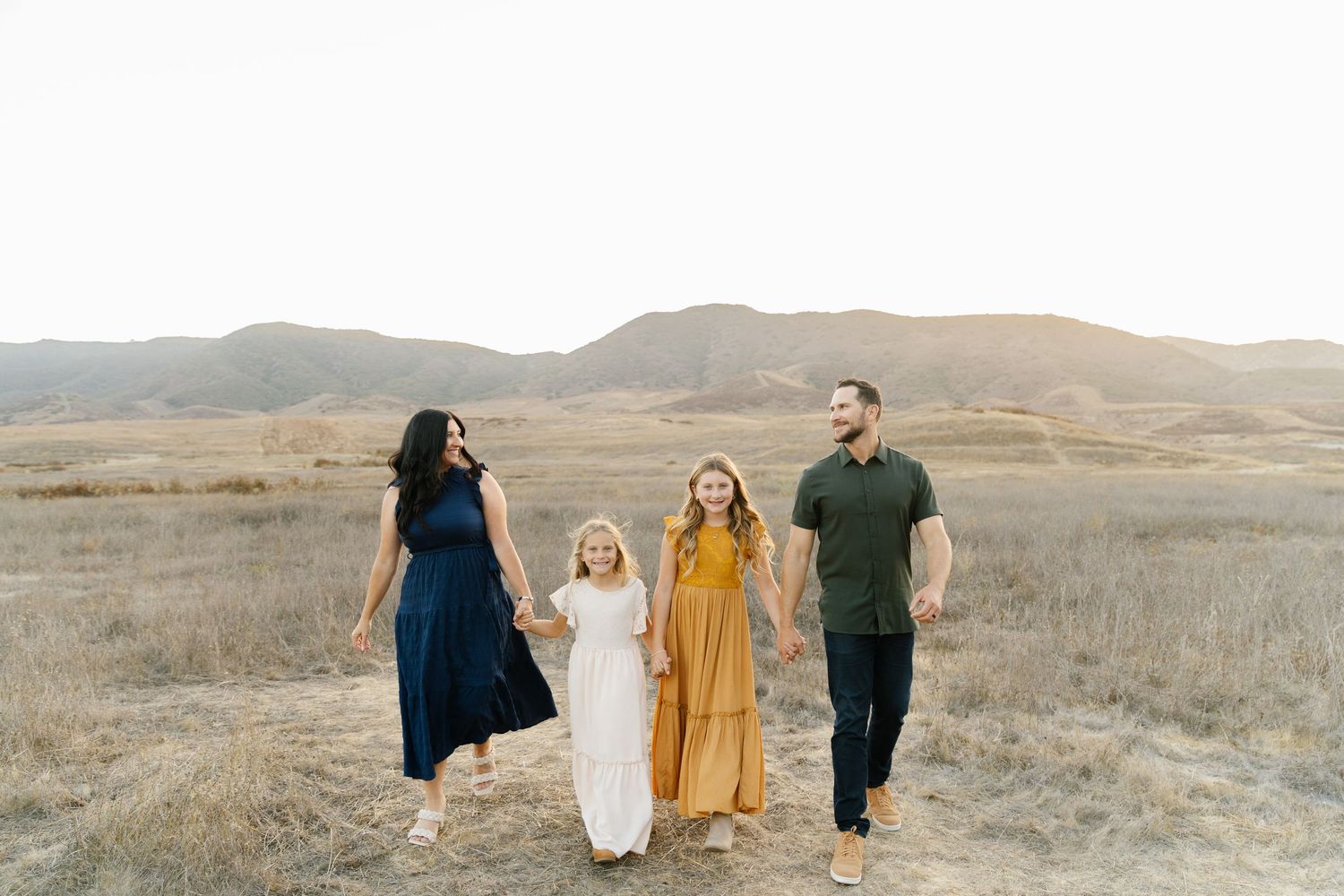 Family of four walking hand-in-hand through golden fields with mountain backdrop during sunset.