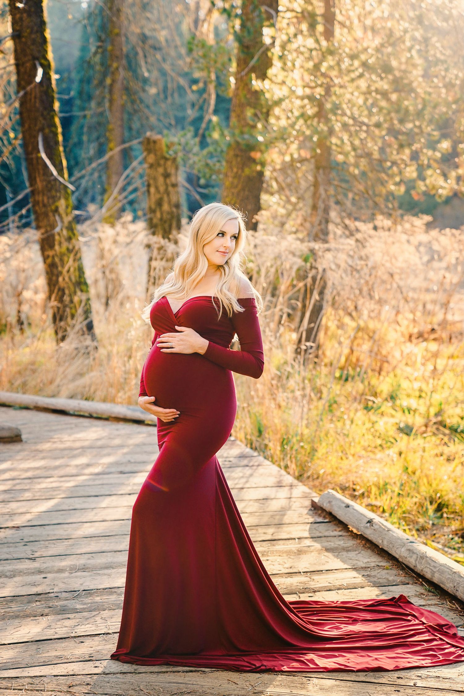 Expecting mother in flowing burgundy gown poses on wooden boardwalk during sunset maternity photoshoot.