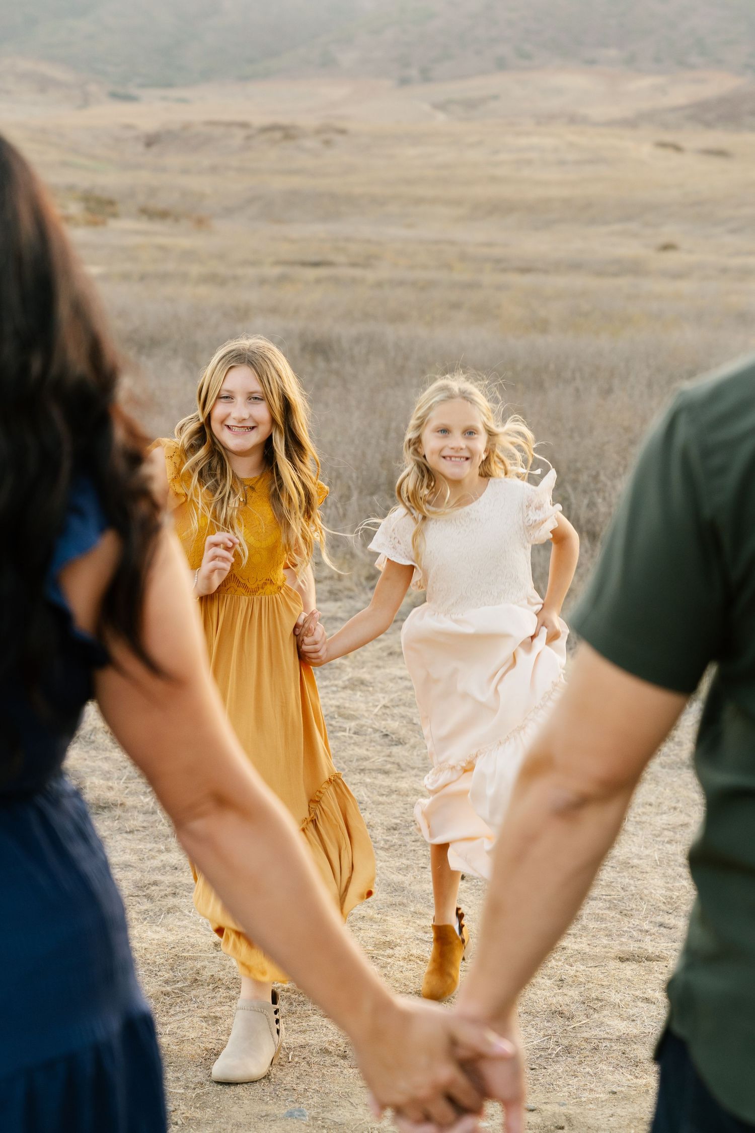 Two smiling girls in yellow and white dresses run toward parents holding hands in a golden field.