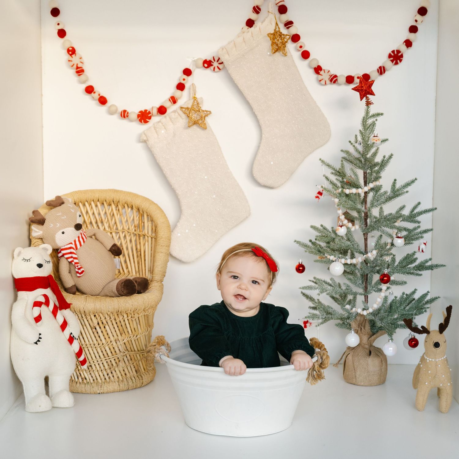 Baby in white tub with Christmas tree, stockings, garland, and stuffed animals in festive holiday photo setting.
