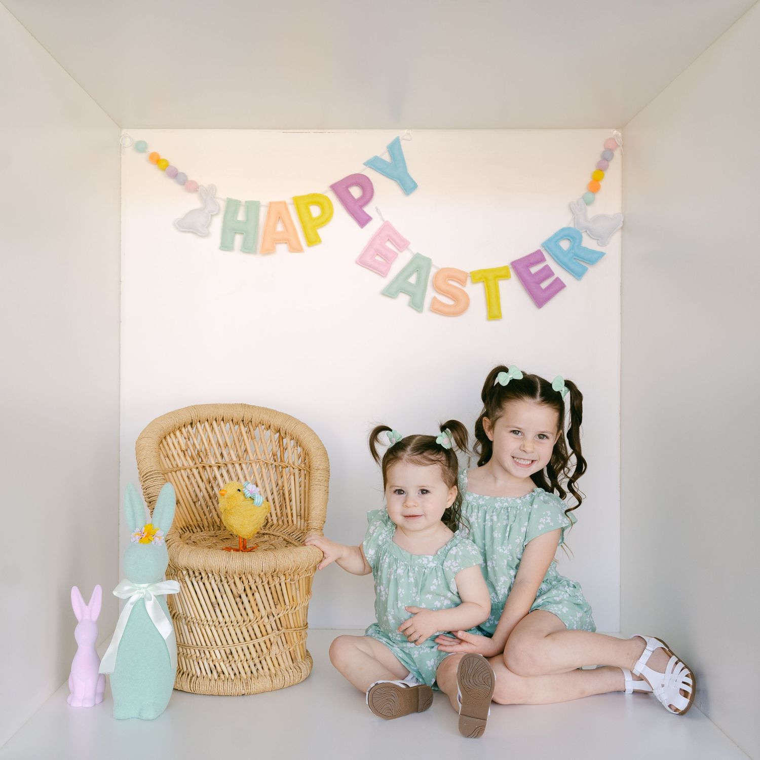 Two small children in matching mint dresses pose with Easter decorations and a wicker chair against a white wall.