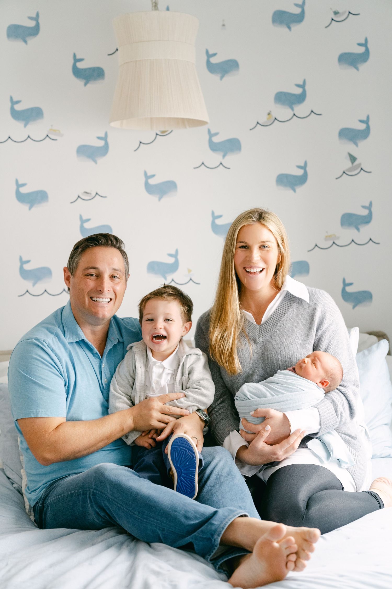 Happy family seated on bed with blue whale pattern wallpaper behind them.