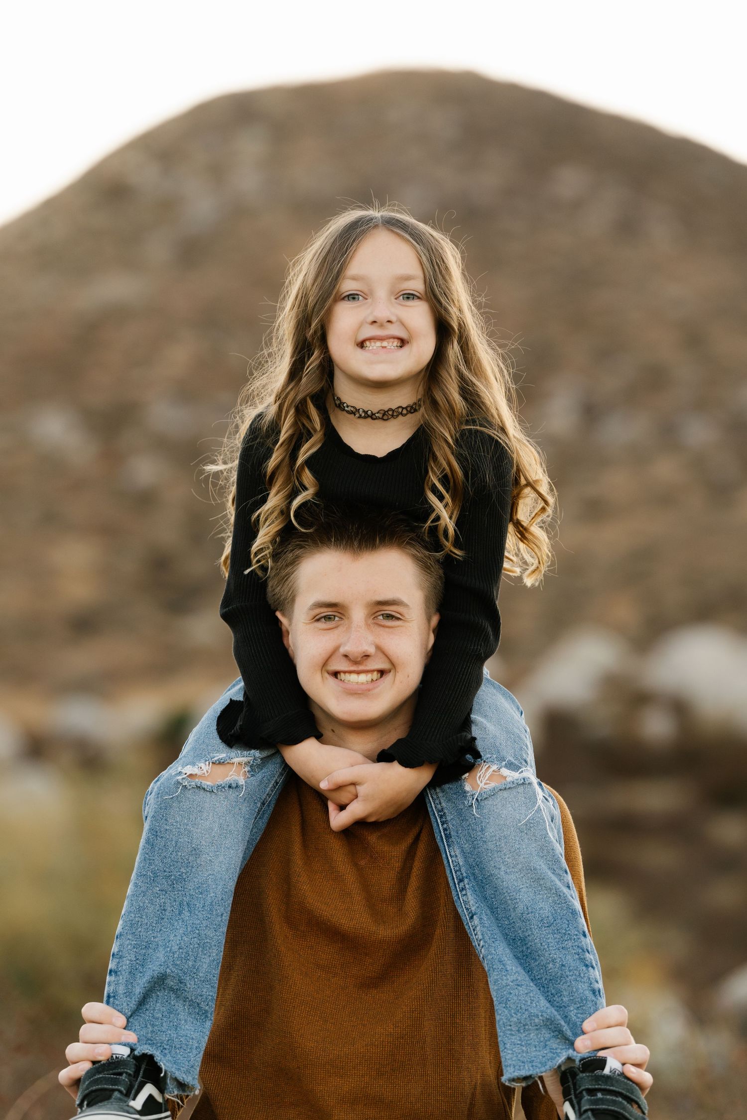 Two young people pose playfully with piggyback ride outdoors against mountainous background at sunset.
