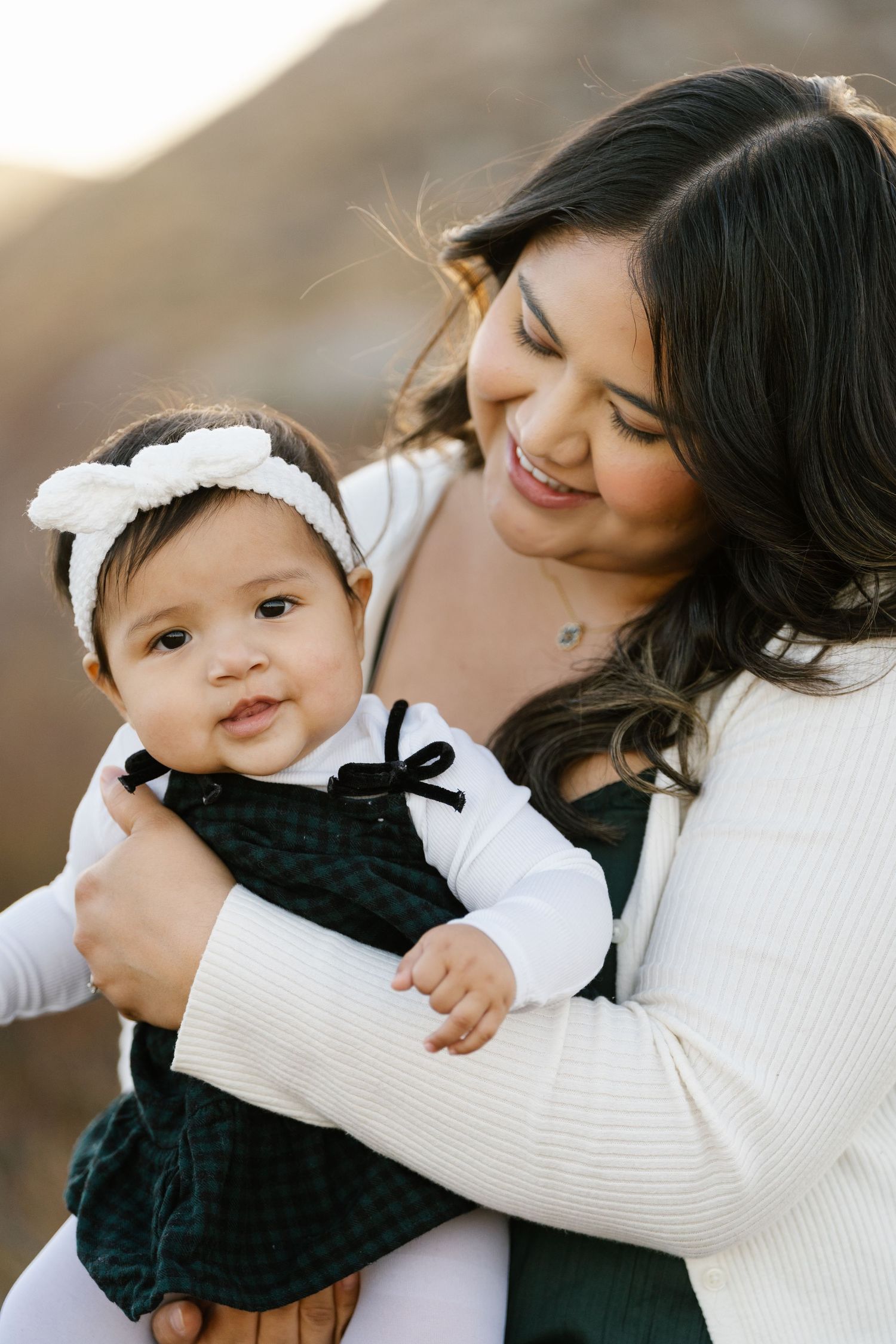 Parent holding baby in velvet dress against mountain backdrop.