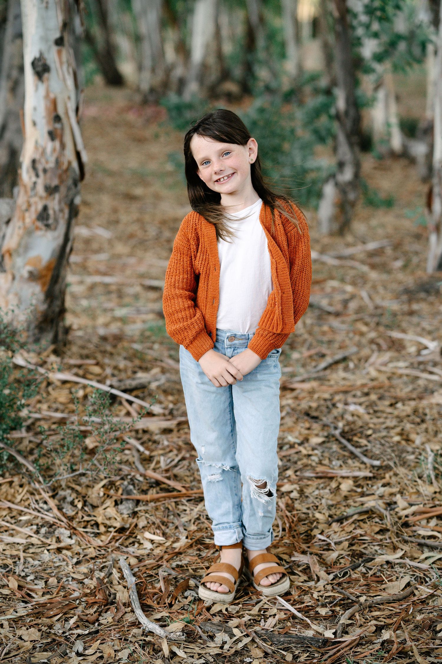 Young girl in orange cardigan, white top, and ripped jeans smiles while standing in a eucalyptus forest.