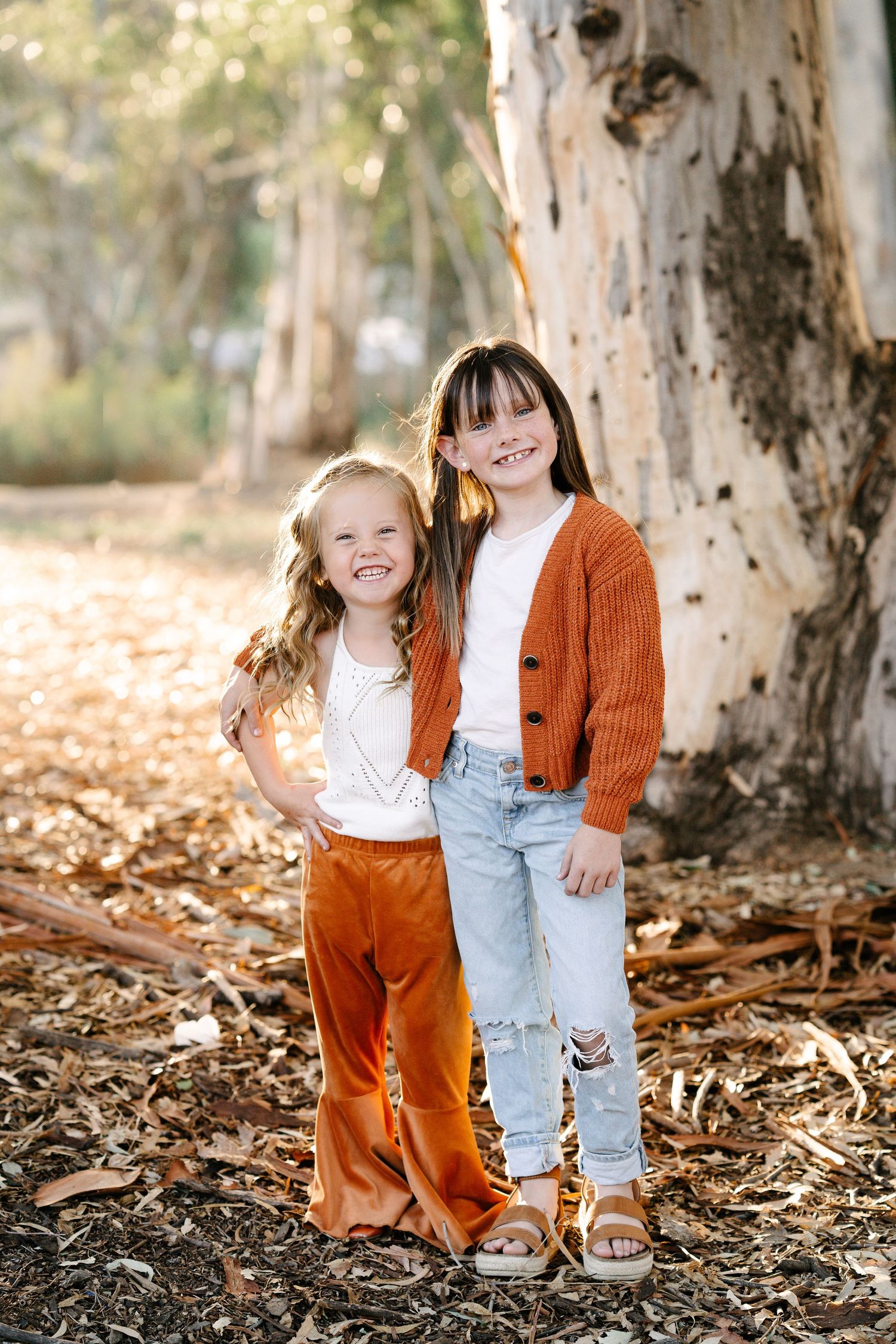 Two young girls in rust orange outfits smile together among fallen leaves in a sunlit eucalyptus forest.