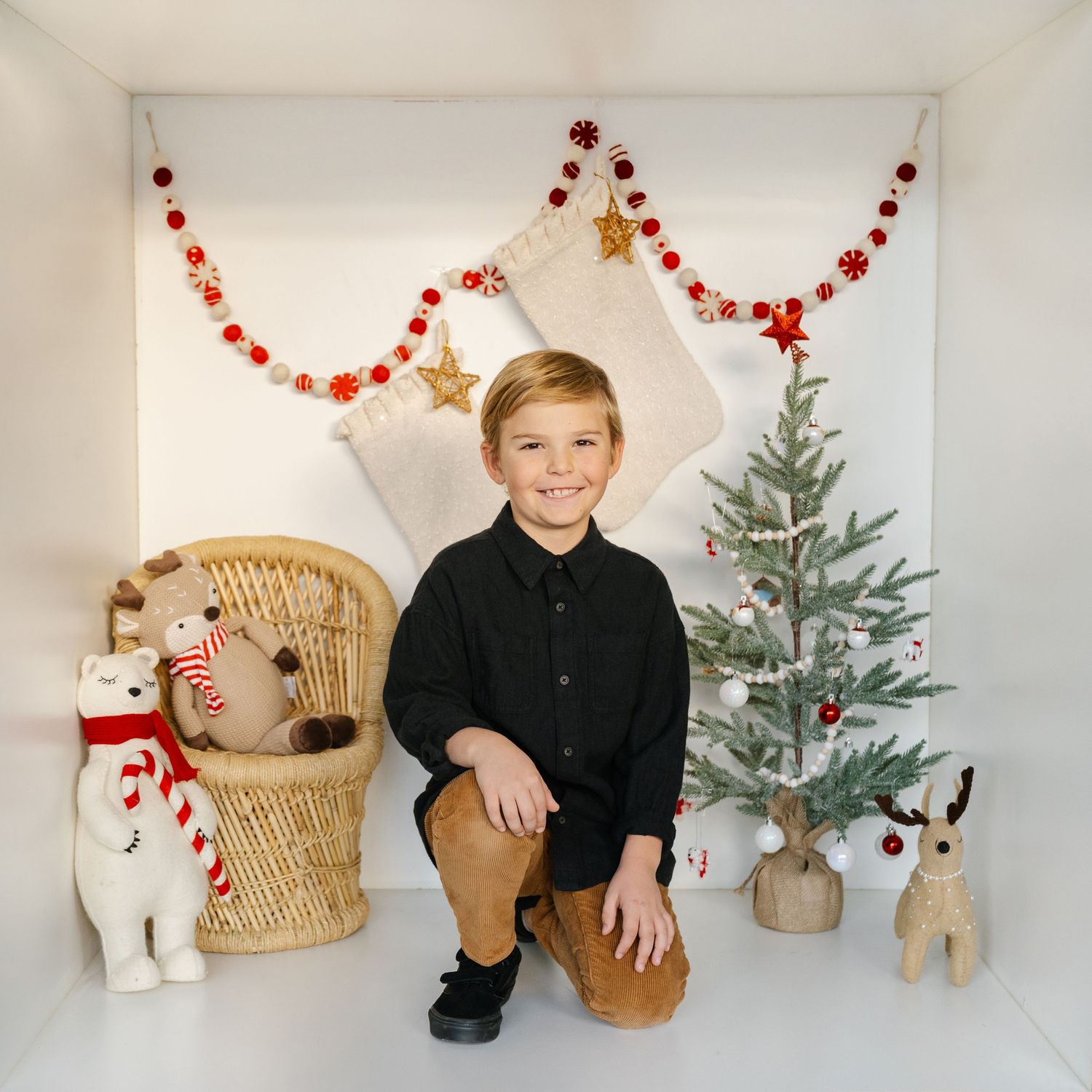 A smiling boy in a black shirt poses in a festive Christmas setting with holiday decorations, stuffed animals, and a small tree.