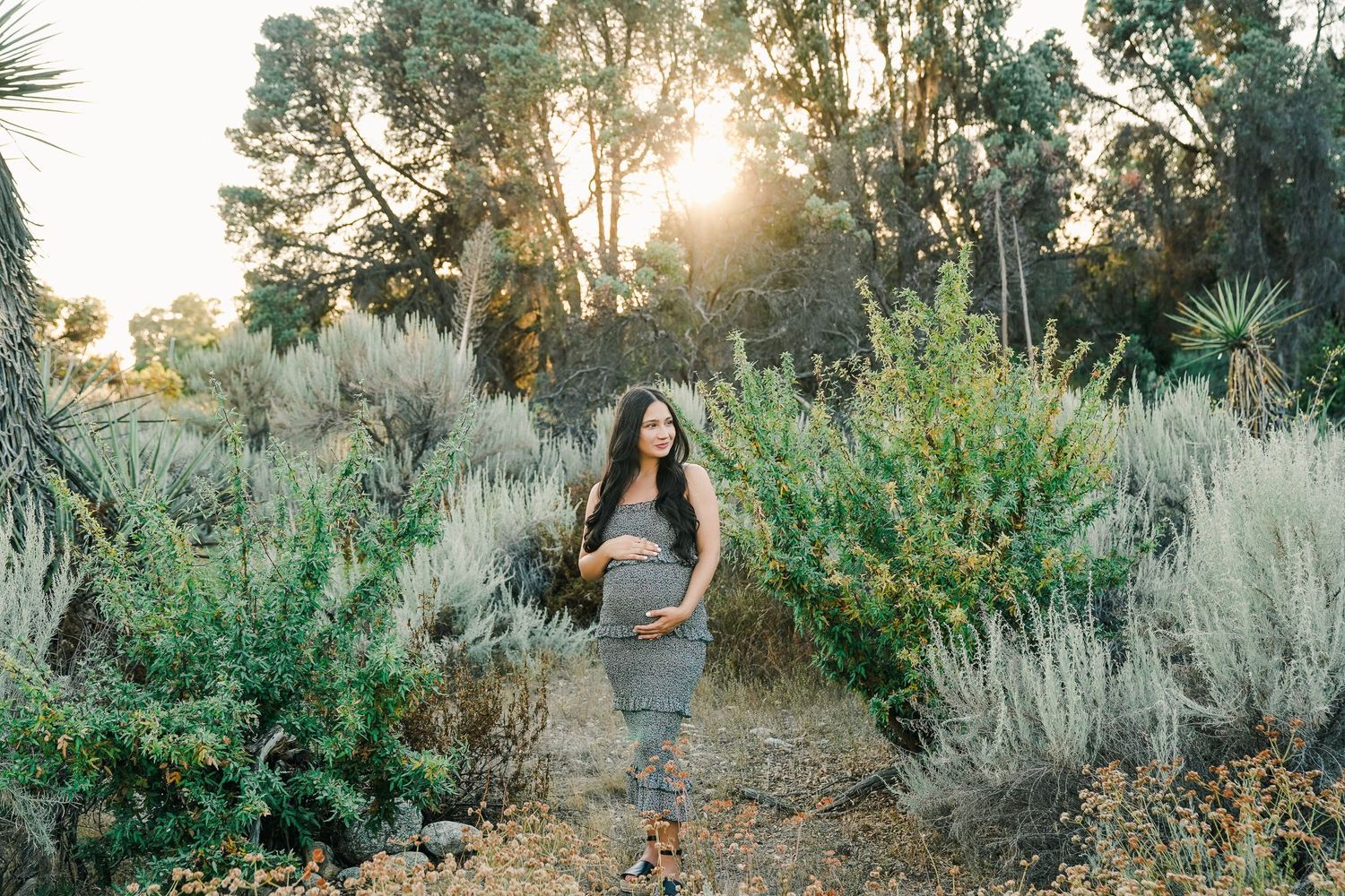 Pregnant woman in grey attire poses among desert brush during golden hour.