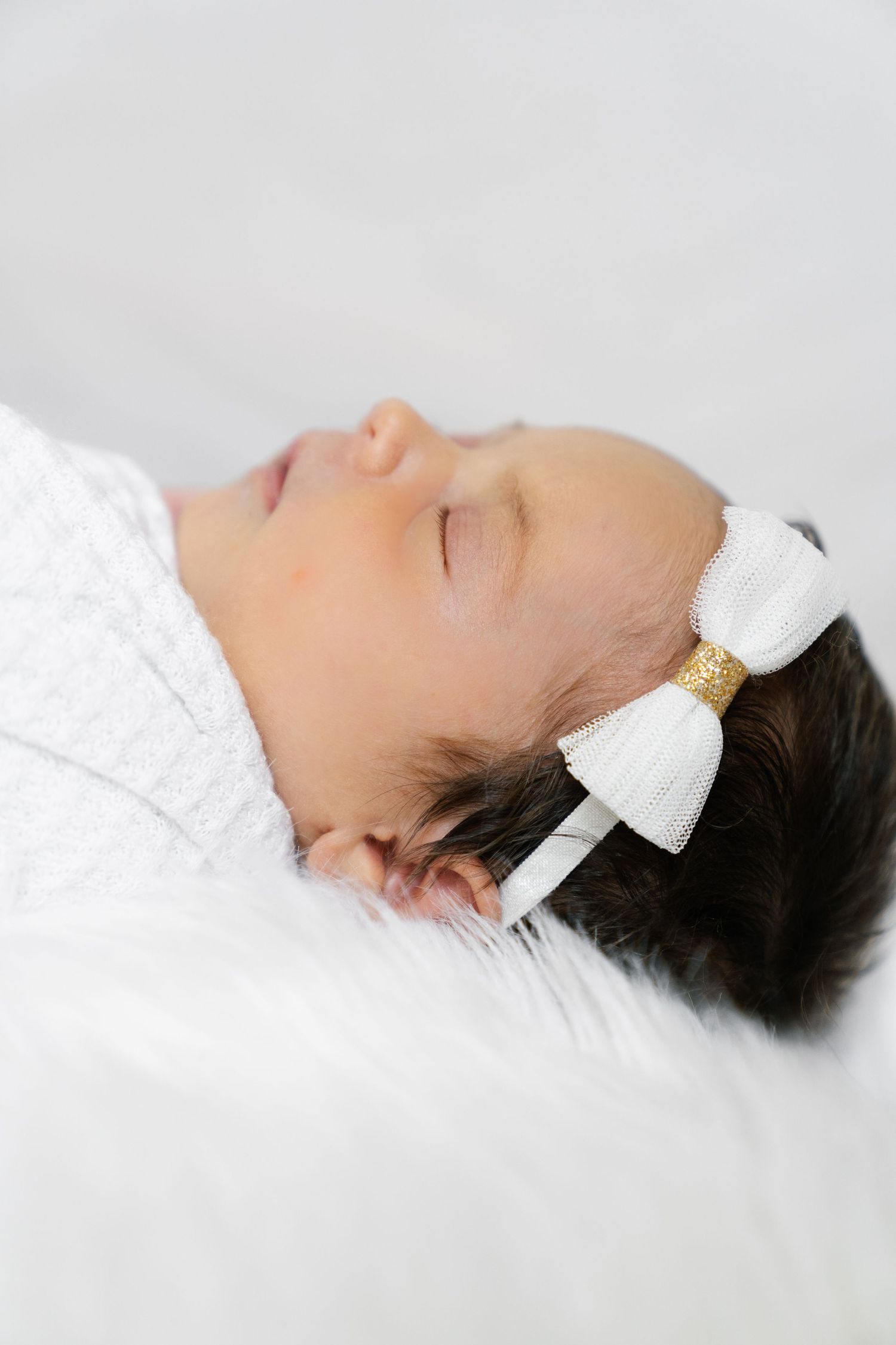Sleeping infant wearing white bow hair accessory on soft white surface.