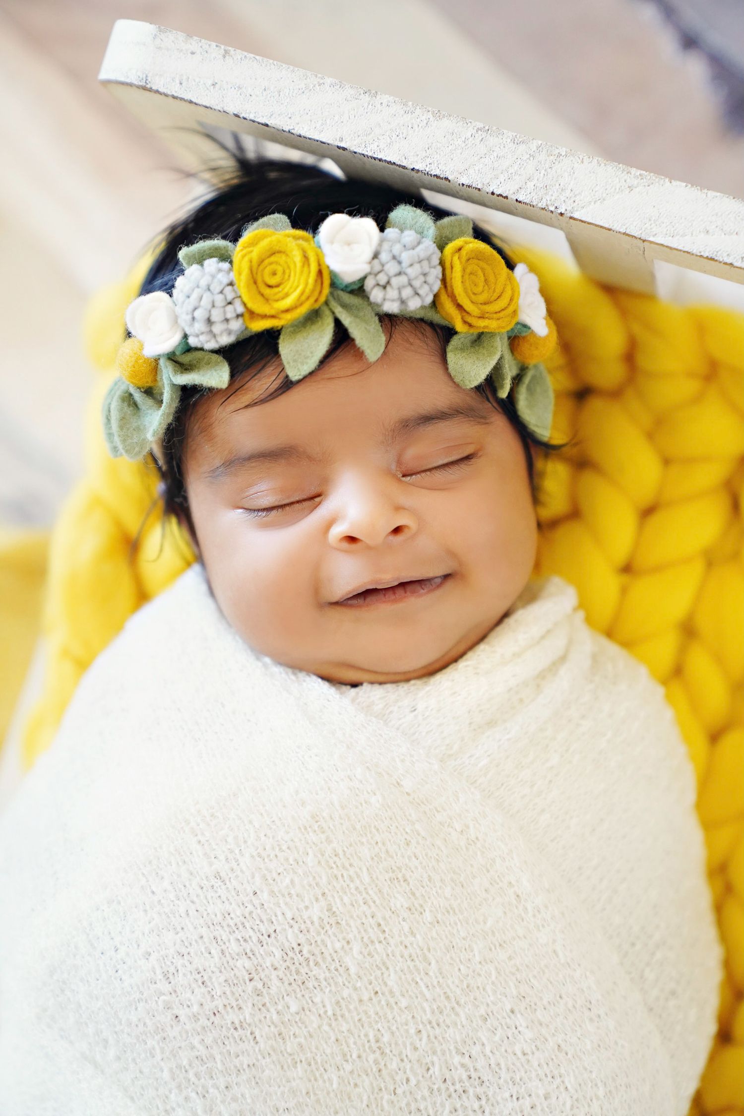 Baby wearing yellow and white floral crown wrapped in white swaddle on yellow blanket.