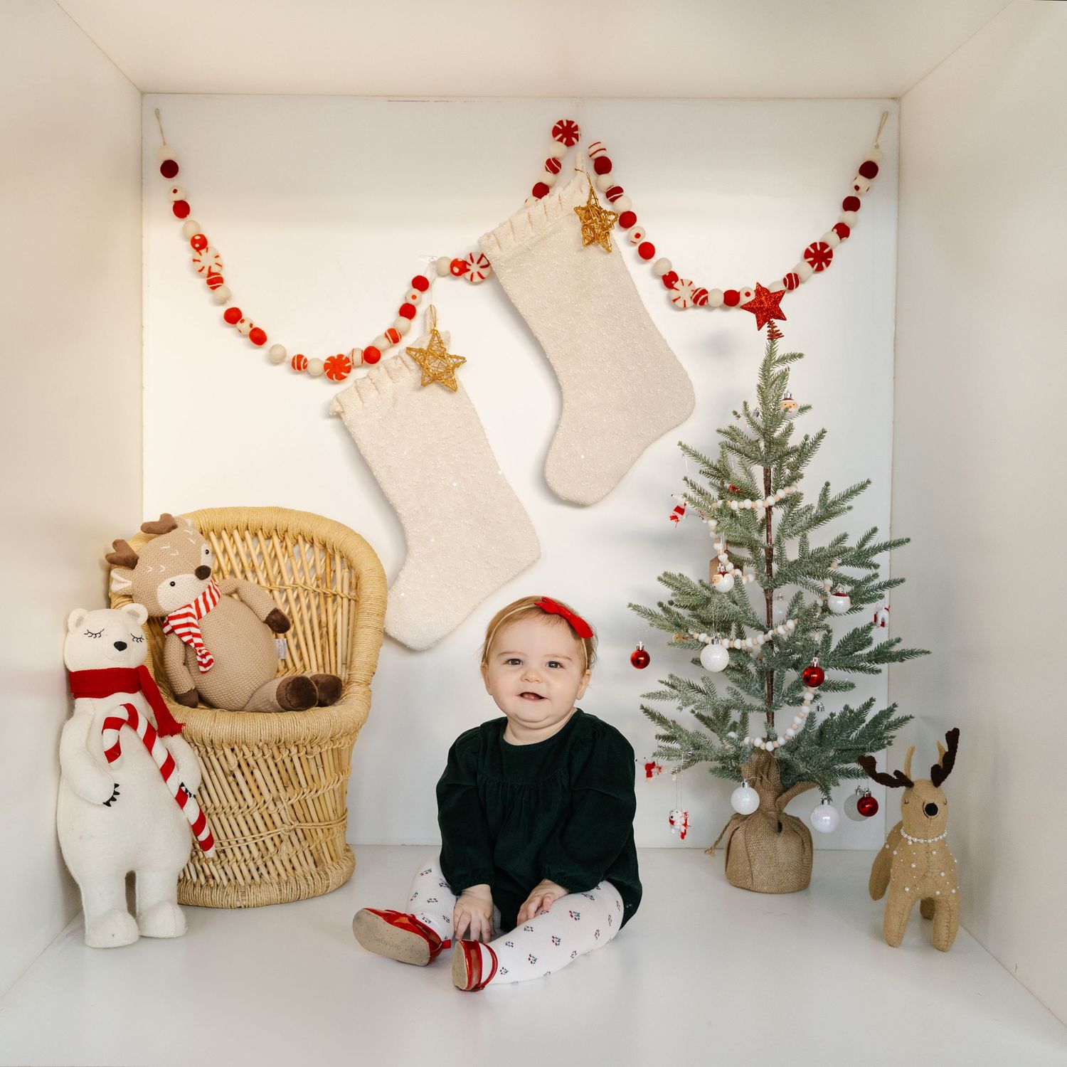 Baby in black outfit sits before Christmas tree, stockings garland, wicker chair, and holiday stuffed animals in white room.