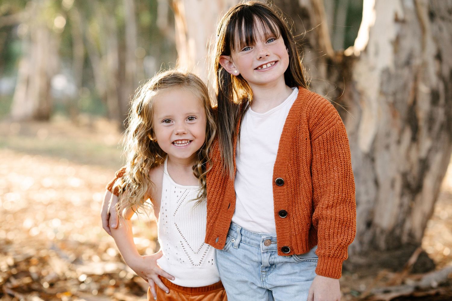 Two smiling young sisters pose together in coordinating rust orange outfits in a sunny woodland setting.