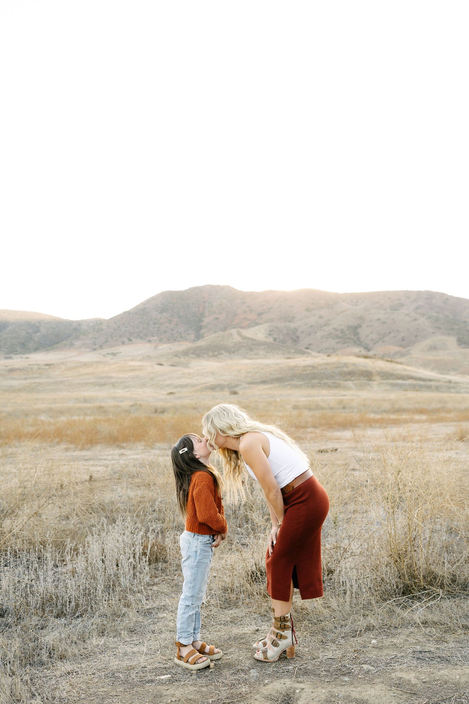 A blonde mother kisses her young daughter in a golden field with rolling hills.