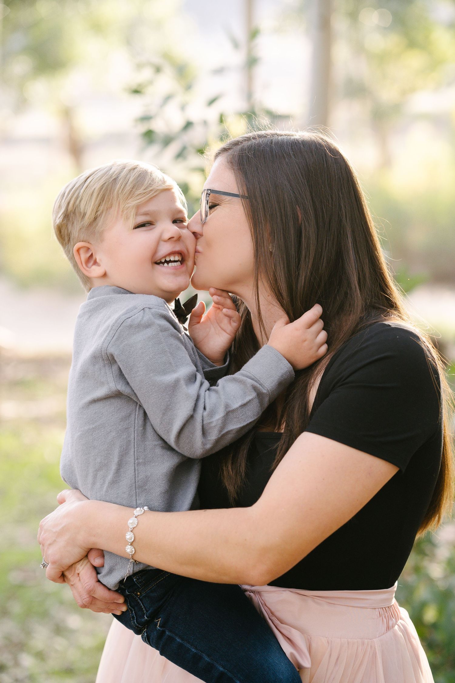A mother kisses her laughing toddler son on the cheek while holding him outdoors in a park.