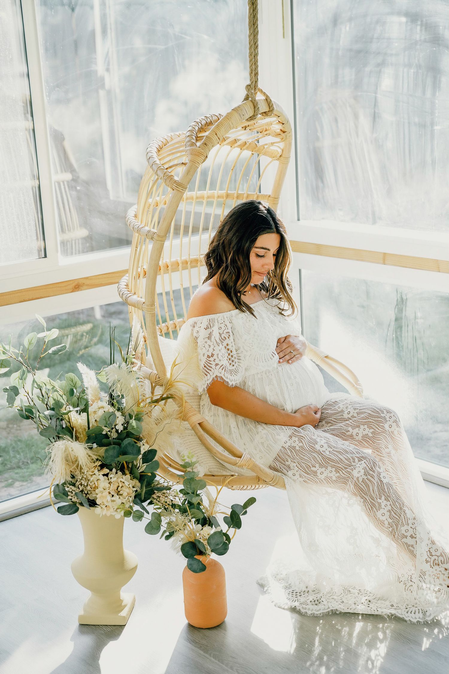 Maternity portrait of woman in lace dress sitting in hanging chair with floral decorations.