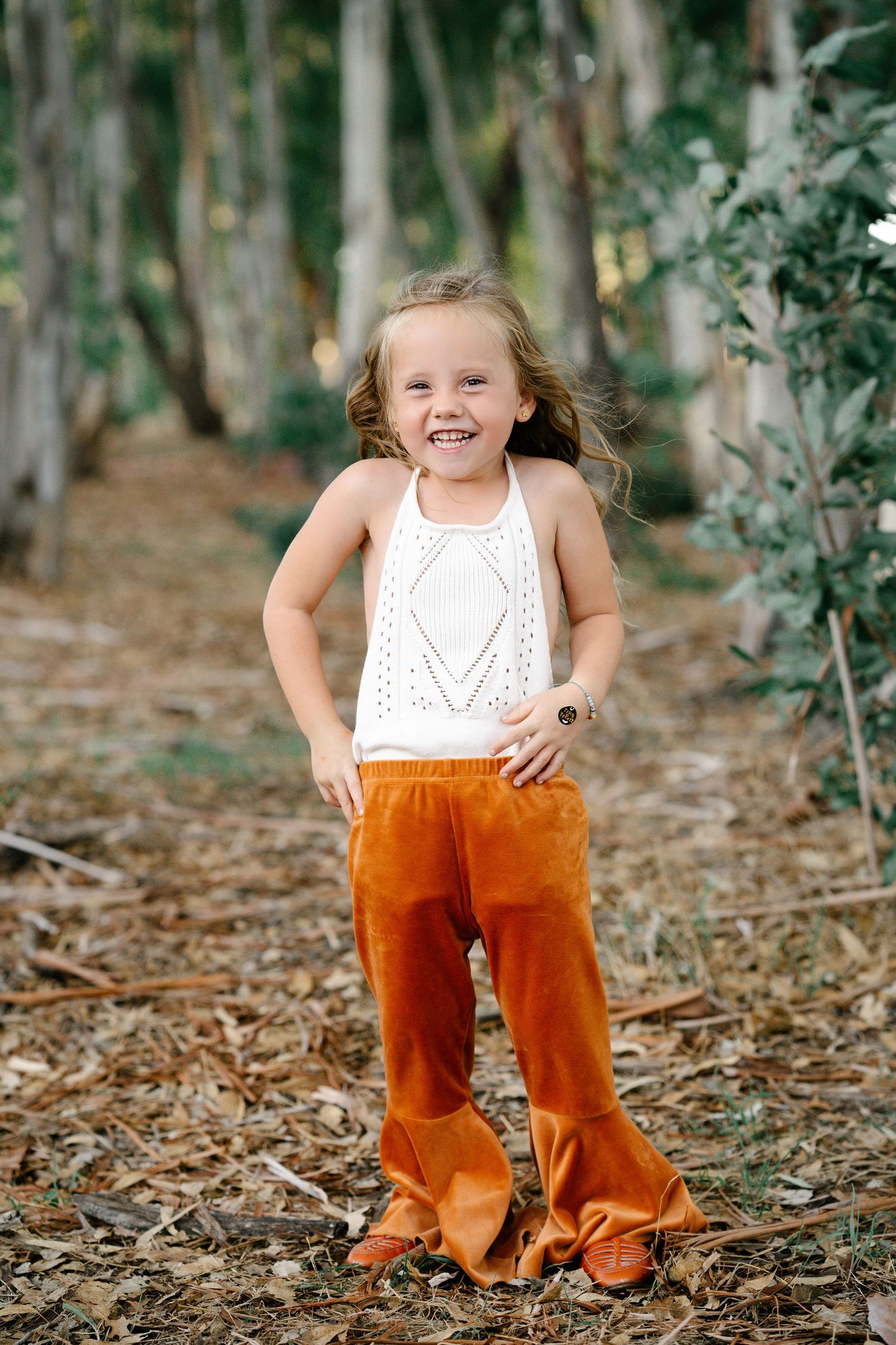 A young girl in orange velvet bell bottoms and white top poses in a forest.