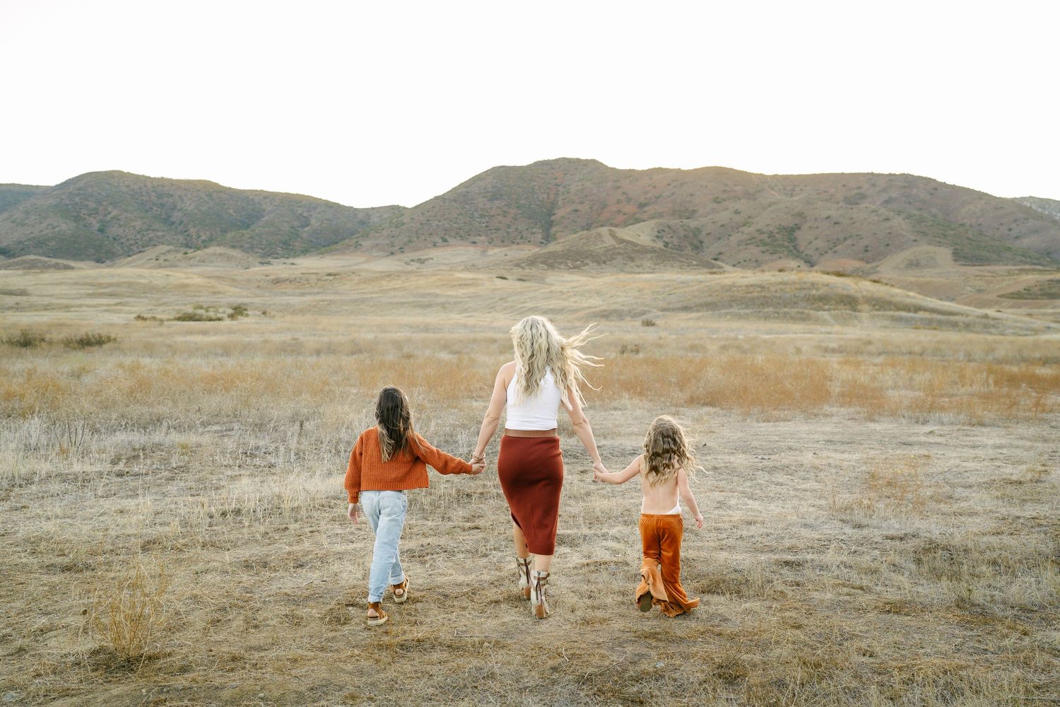 A mother walks hand-in-hand with two young children through a golden field with rolling hills in the background.