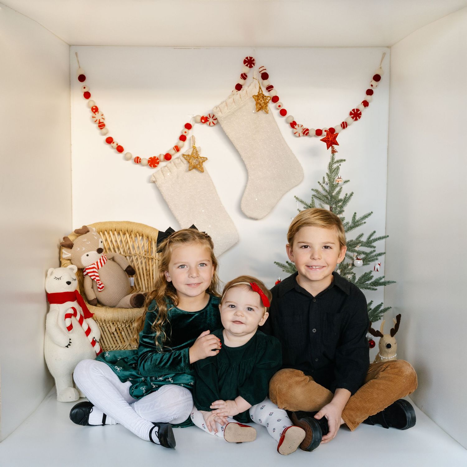 Three children pose together in a festive Christmas photo with stockings, garland, and a small tree in the background.