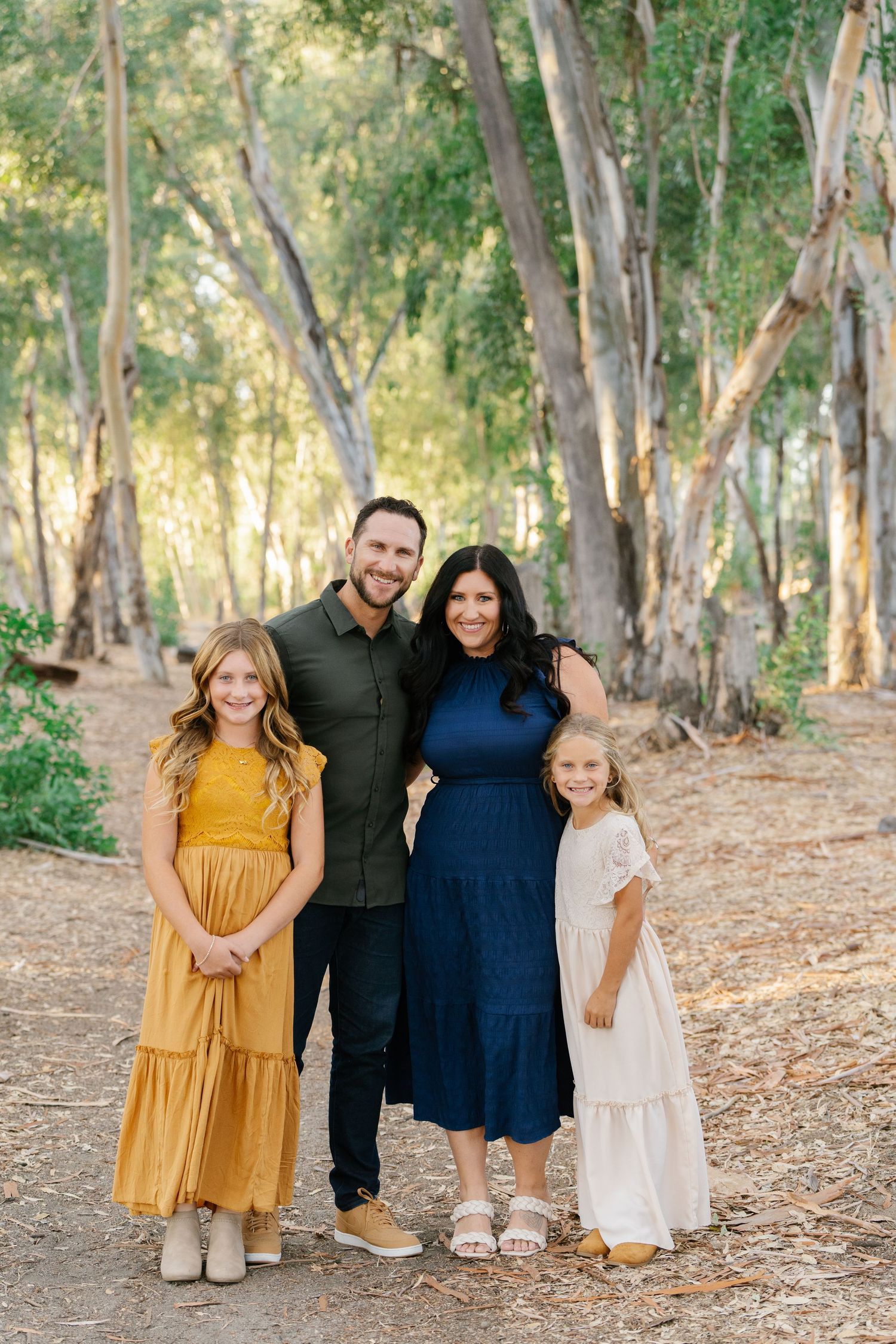 Family of four posing in a sunlit eucalyptus forest, wearing navy, gold, and white outfits.
