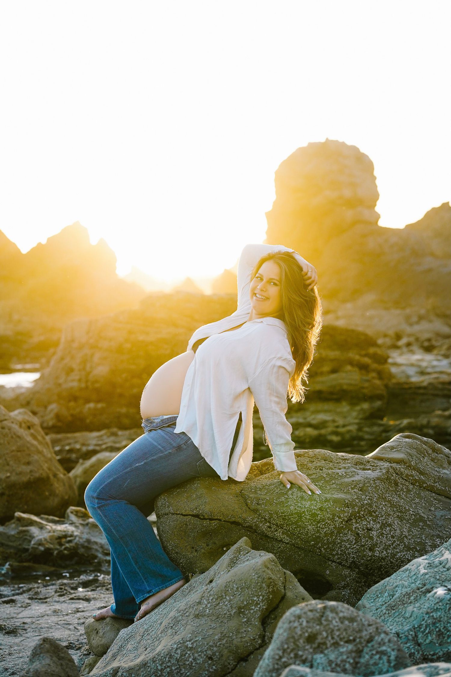 Expectant mother in white top and jeans poses on coastal rocks during golden hour.