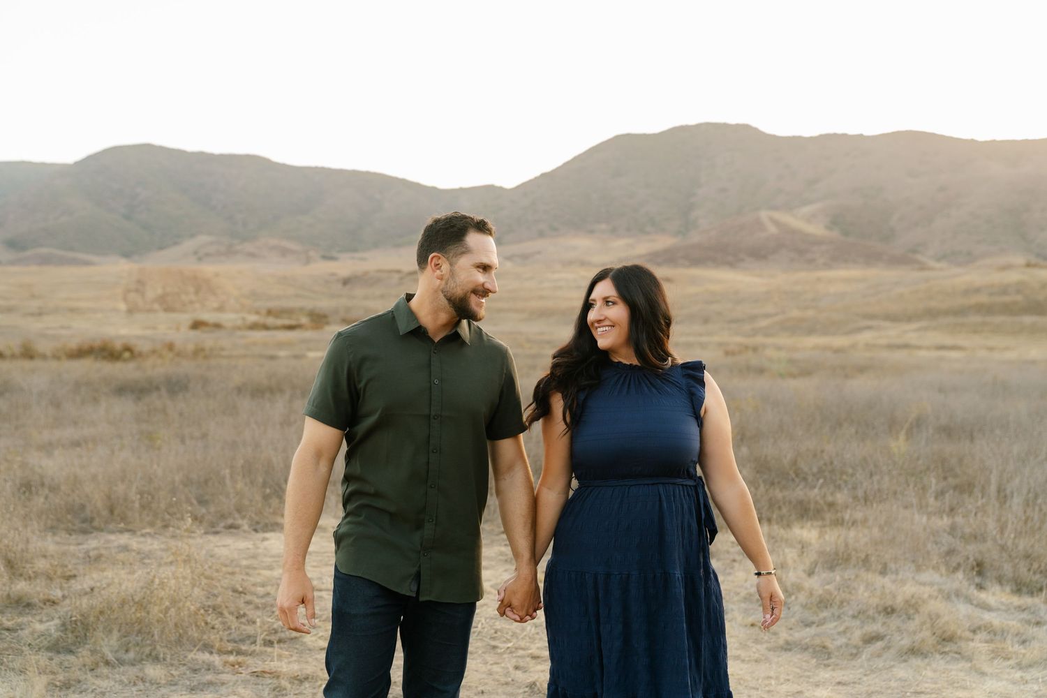 A couple holds hands and smiles while walking through a golden field with rolling hills at sunset.