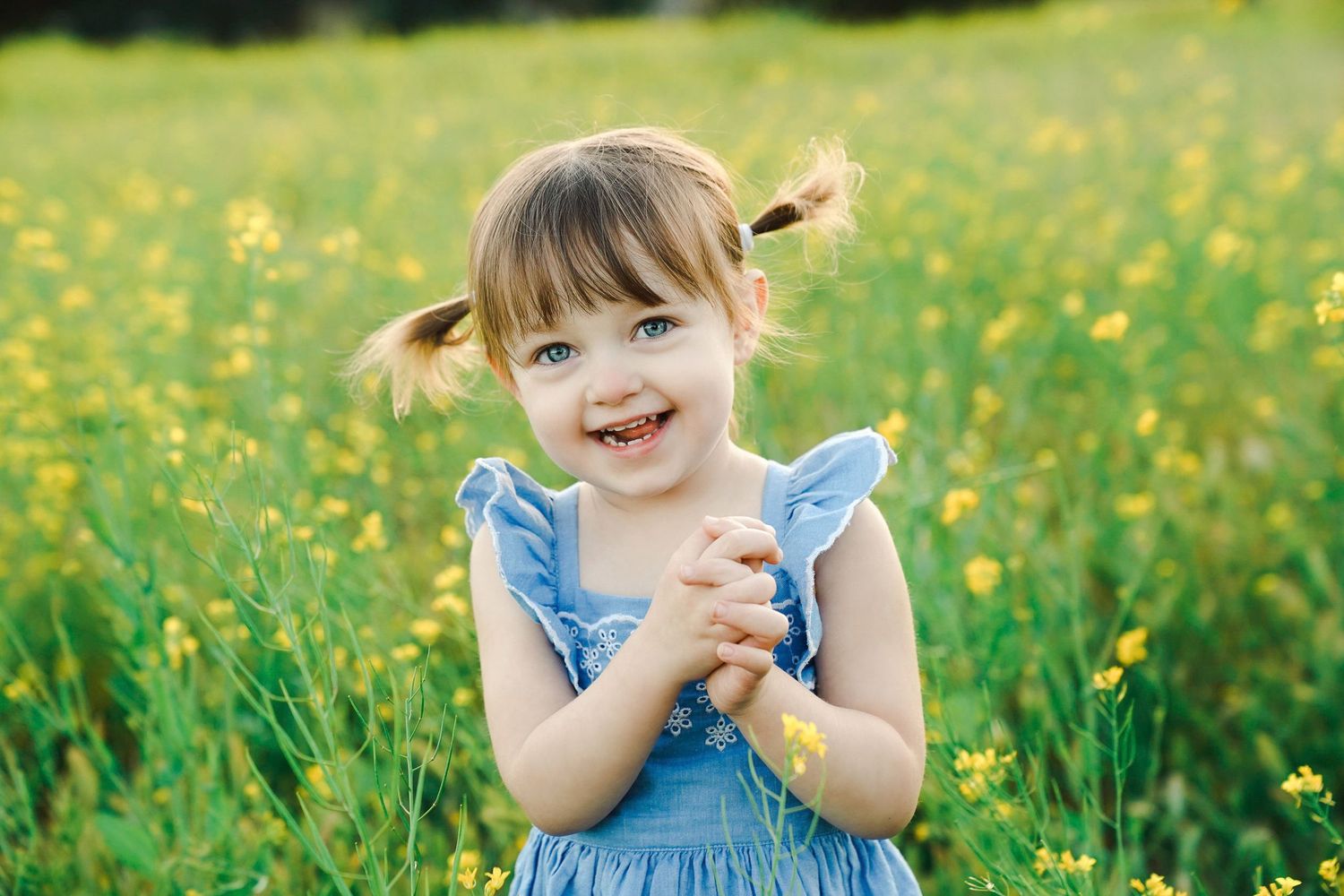 Smiling child in blue dress standing in field of yellow wildflowers.