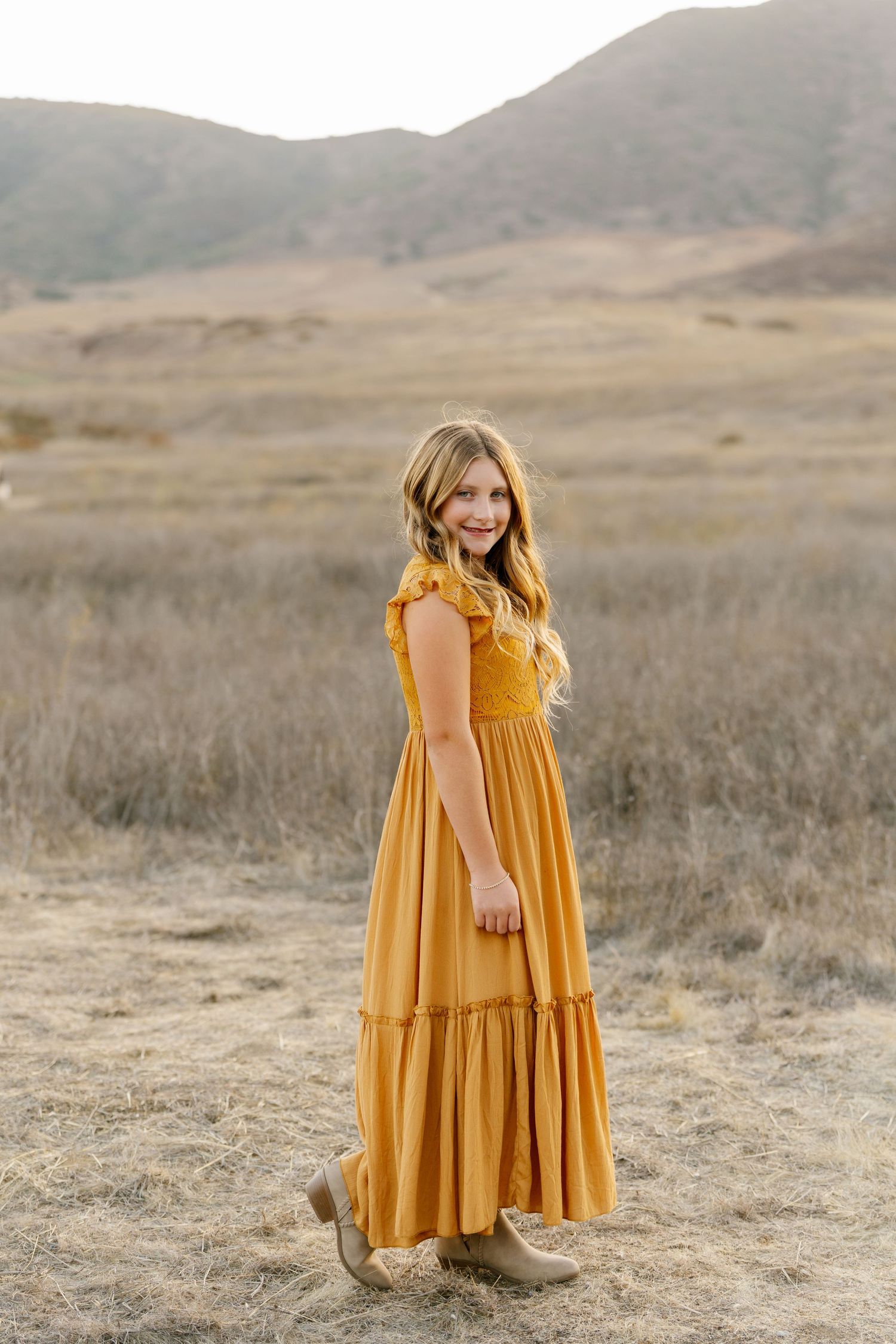A smiling girl in a mustard yellow maxi dress and tan boots stands in a dry field with mountains behind her.
