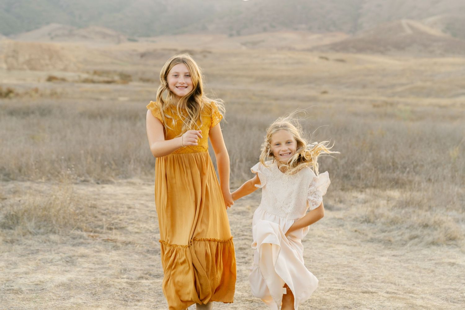 Two women in flowing dresses, one gold and one white, joyfully running through a sunlit golden field.