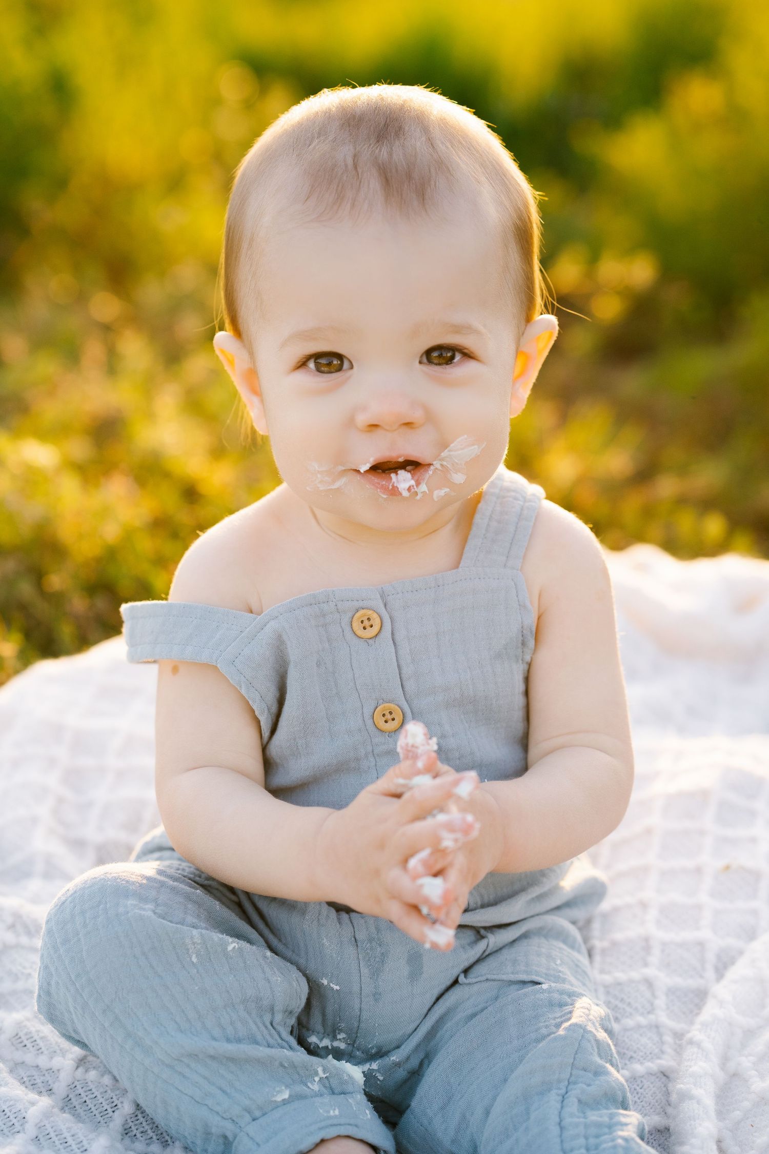 Baby in blue overalls sits on white blanket with golden sunset lighting in background.