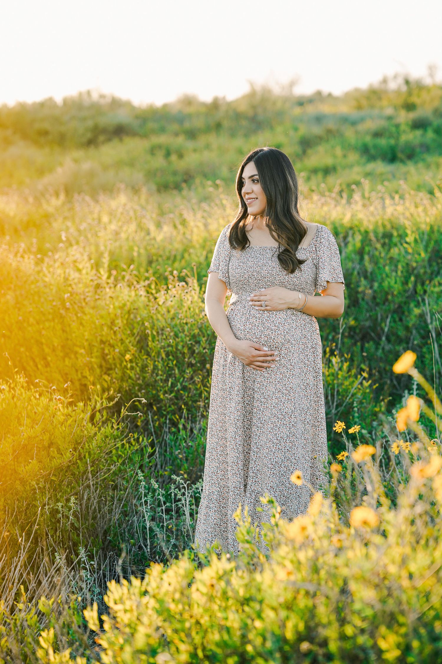 Sunset maternity portrait in lace dress surrounded by golden wildflowers in meadow.