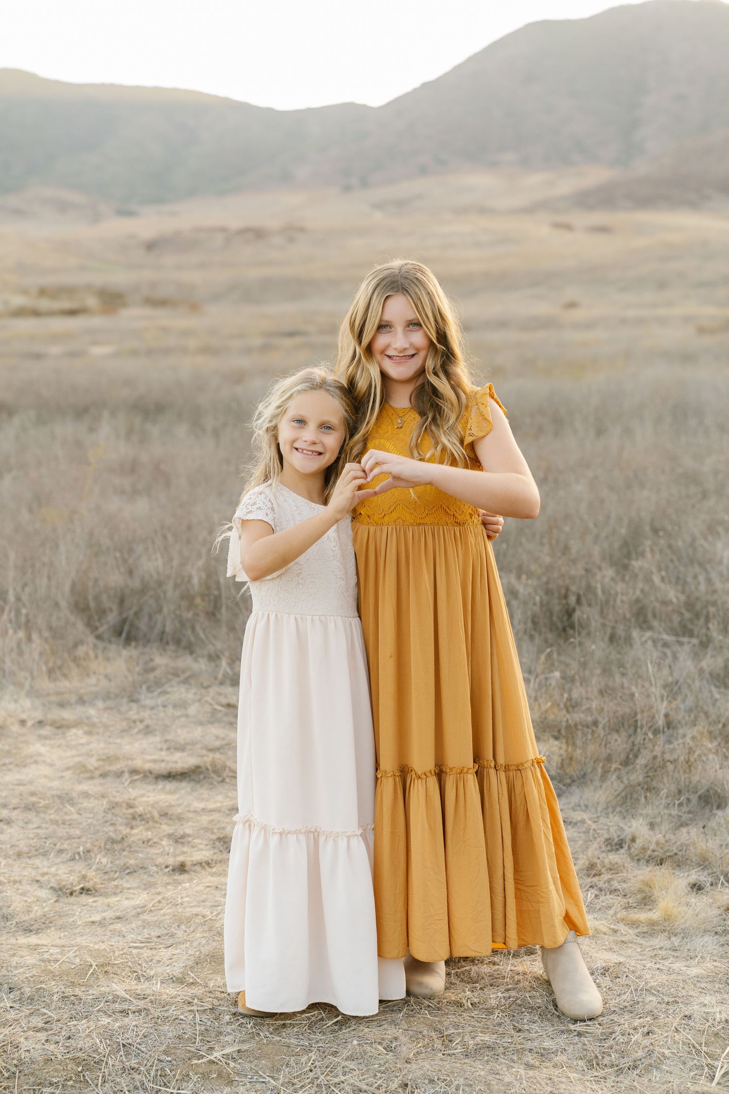 Two young sisters in white and mustard maxi dresses pose together in a golden field with mountains behind.