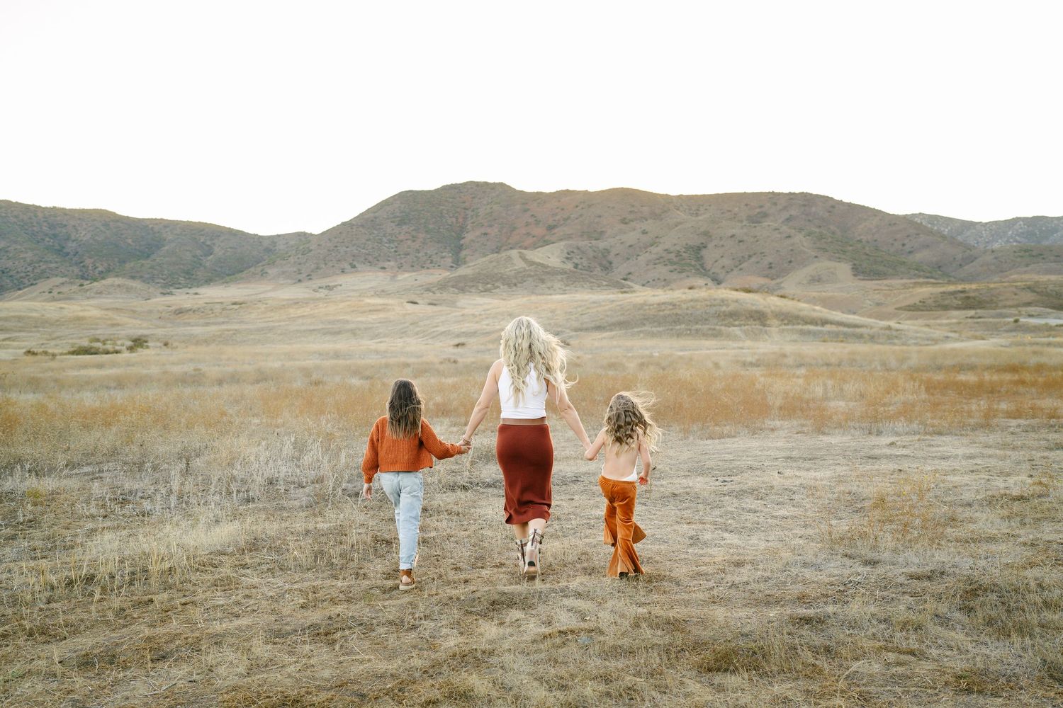 A mother and two children walk hand-in-hand through a golden field with rolling hills in the background.