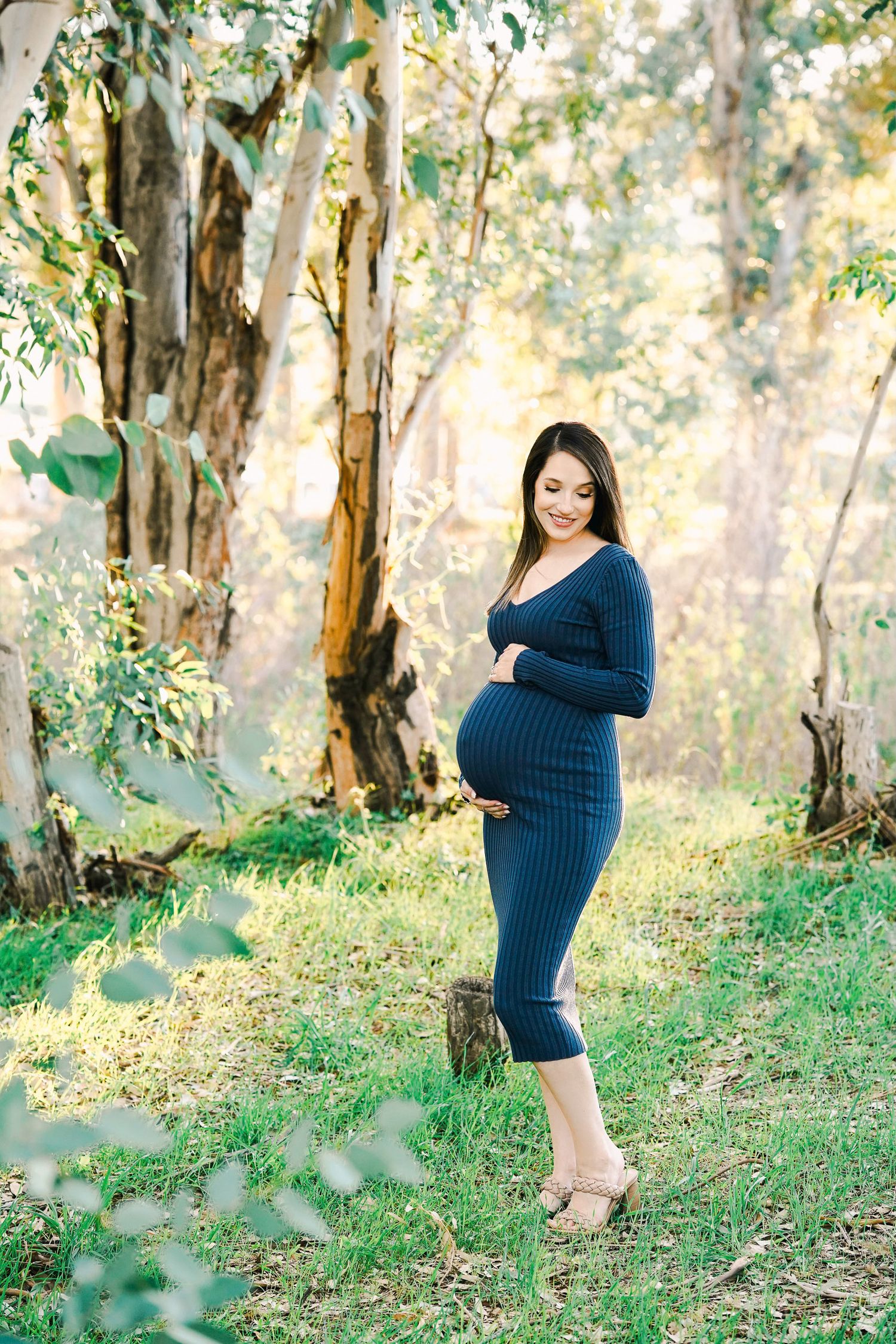 Expectant mother in navy blue dress poses elegantly in sunlit woodland setting.